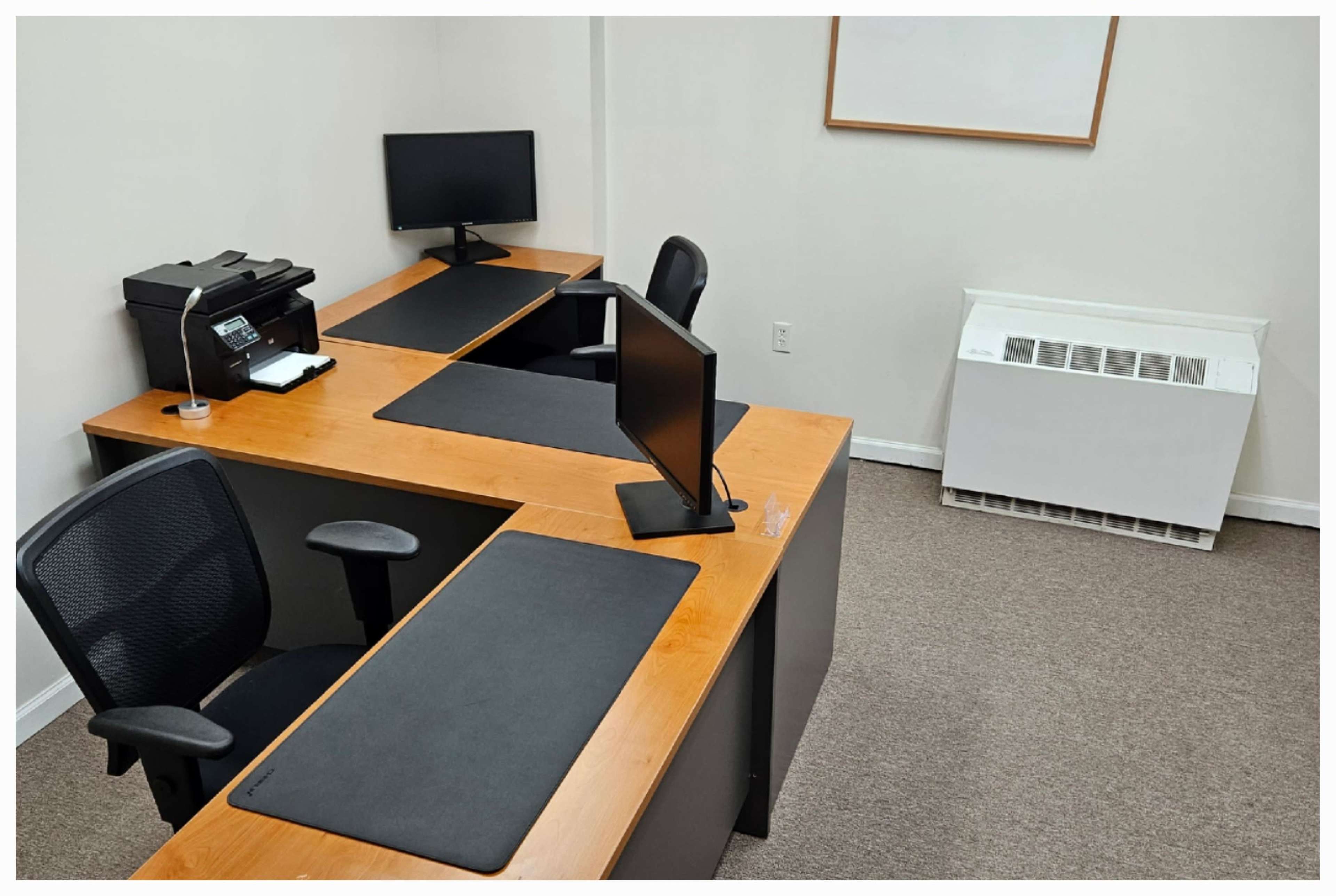 A small office space featuring two desks with computer monitors, a printer, and a heating unit along the wall.