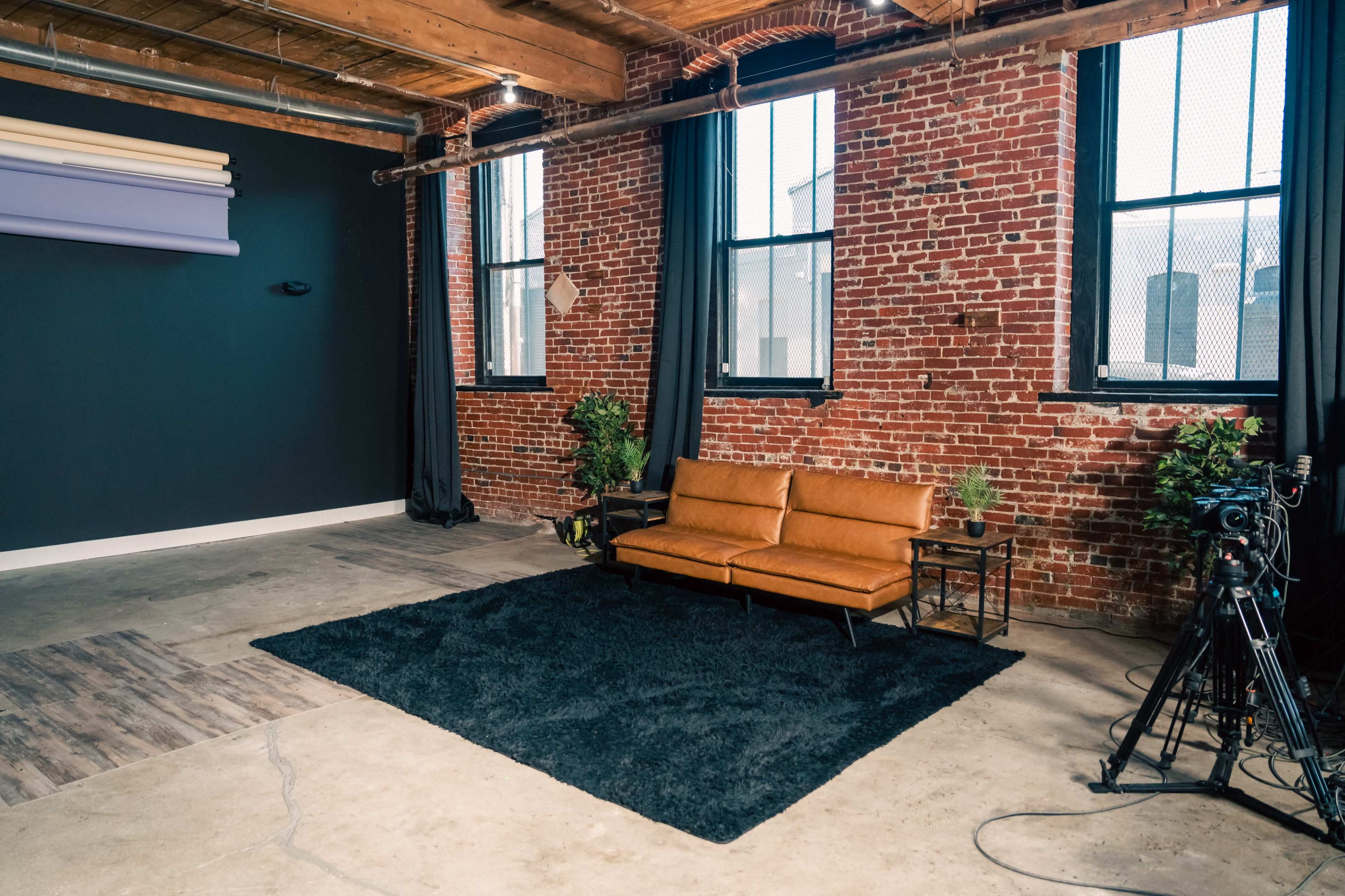 The image shows a minimalistic studio space featuring a brown leather sofa, a black rug, potted plants, and a camera setup near large windows with exposed brick walls.