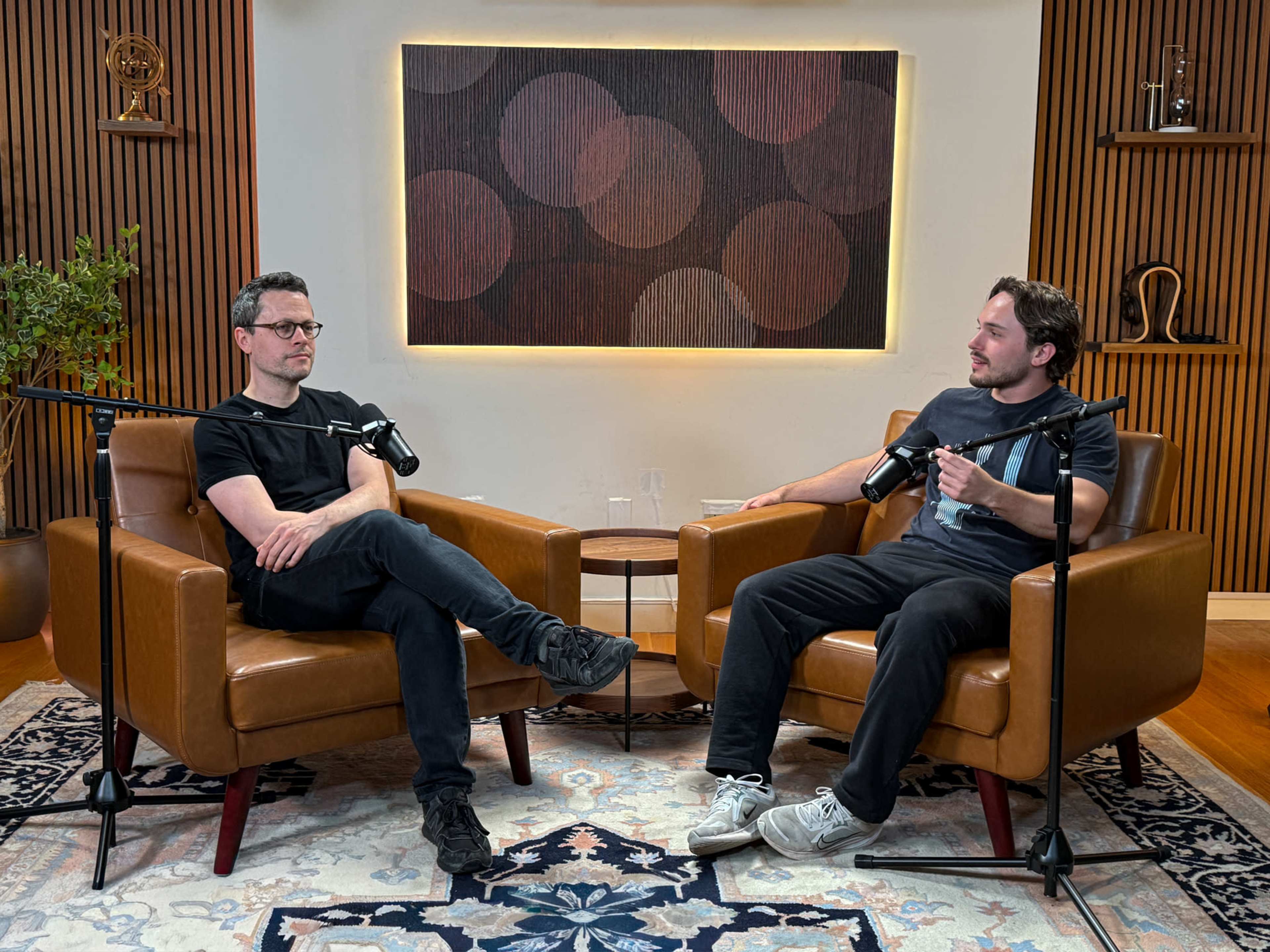 Two men are seated in brown leather armchairs in a modern studio setting, engaged in conversation with microphones in front of them.