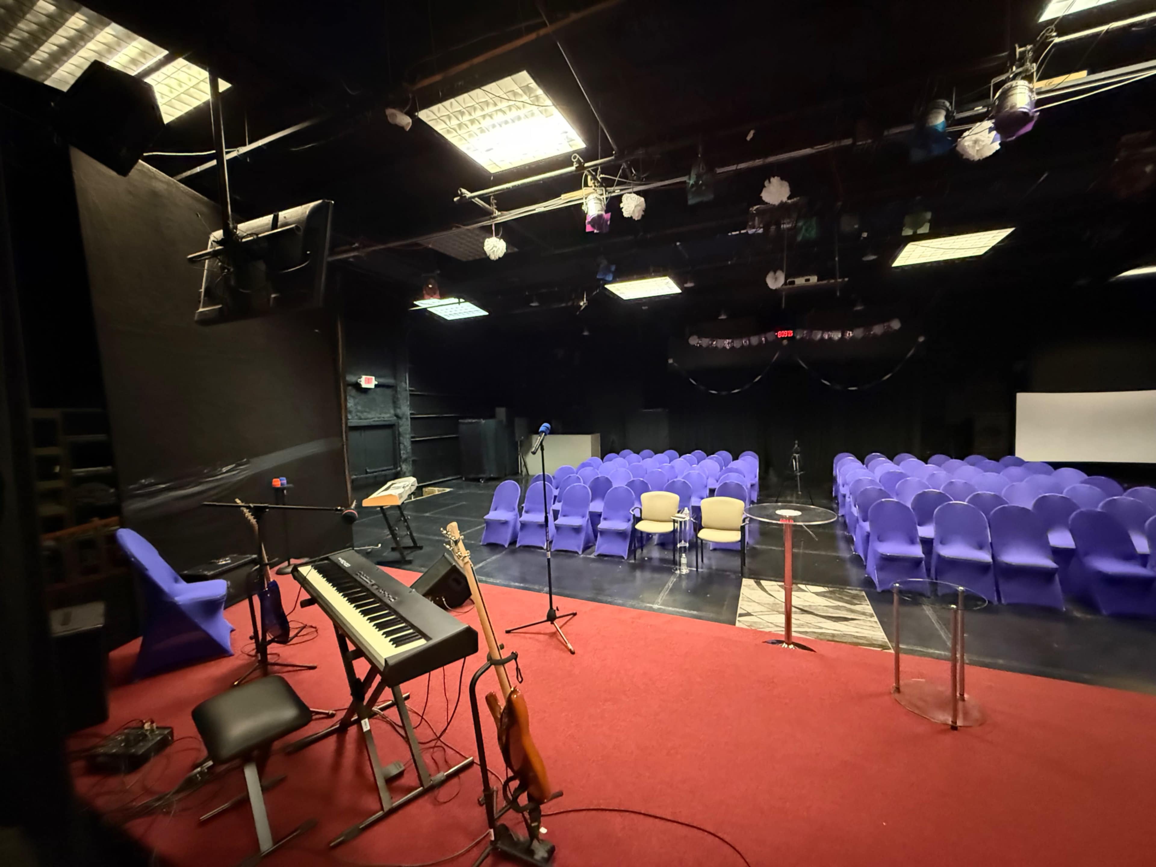 The image shows a dimly lit auditorium with purple chairs arranged for an audience and a stage featuring a keyboard and a microphone setup.