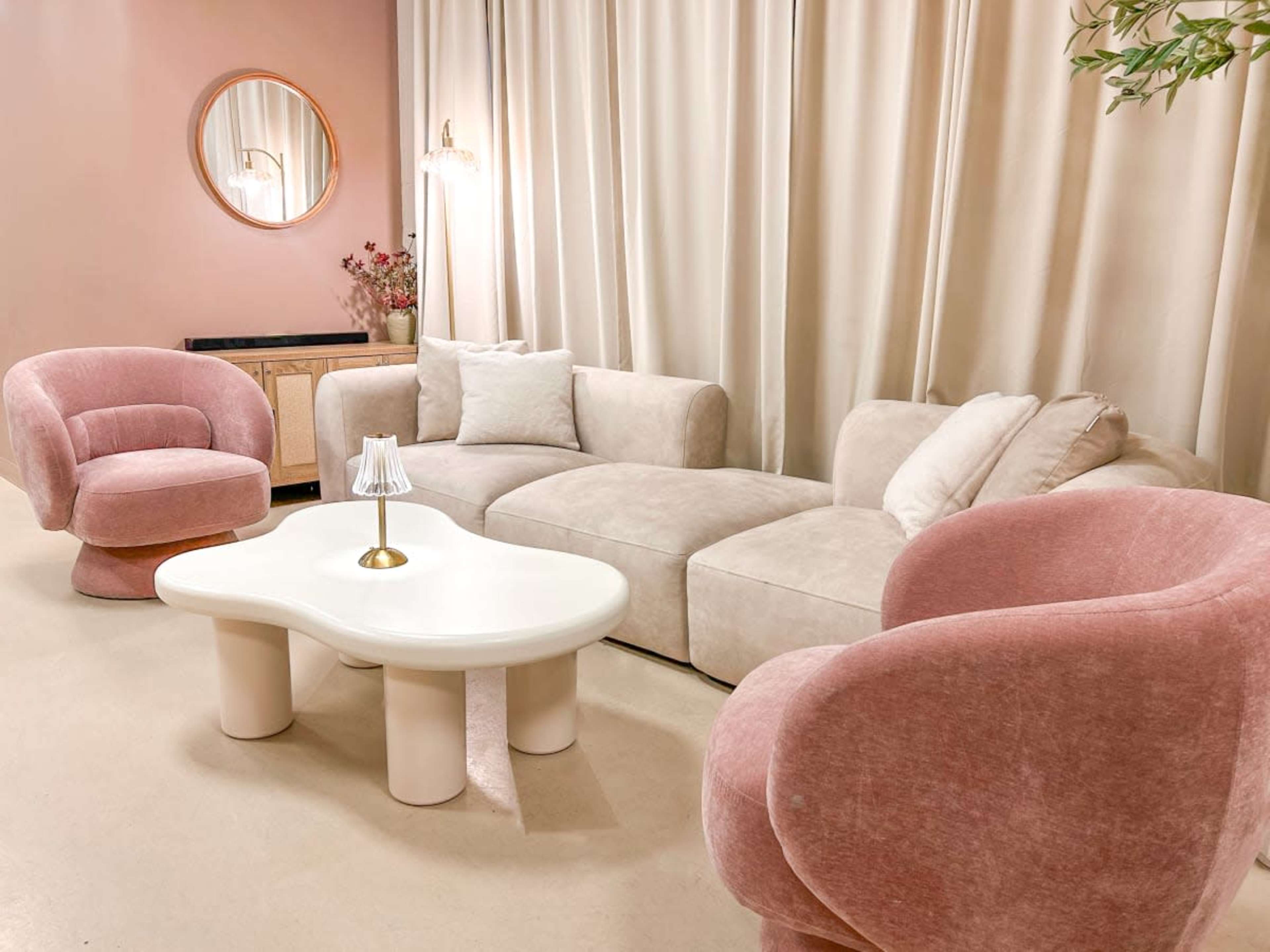 The image features a modern living room with a light pink and beige color scheme, showcasing a curved sofa, two accent chairs, and a round coffee table.