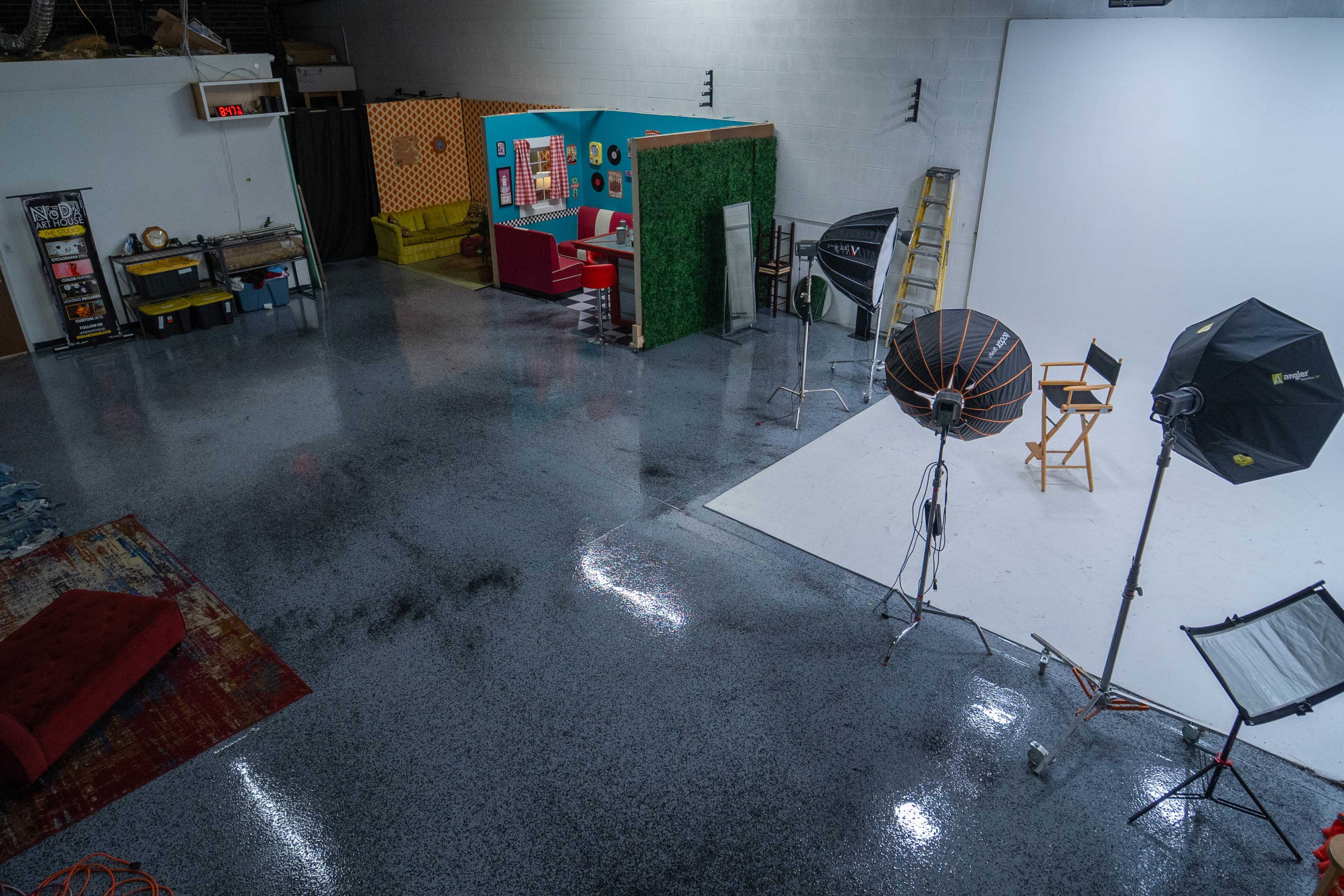 The image shows a spacious studio with a polished floor, various lighting equipment, and distinct seating areas, including a red couch and decorative backdrops.