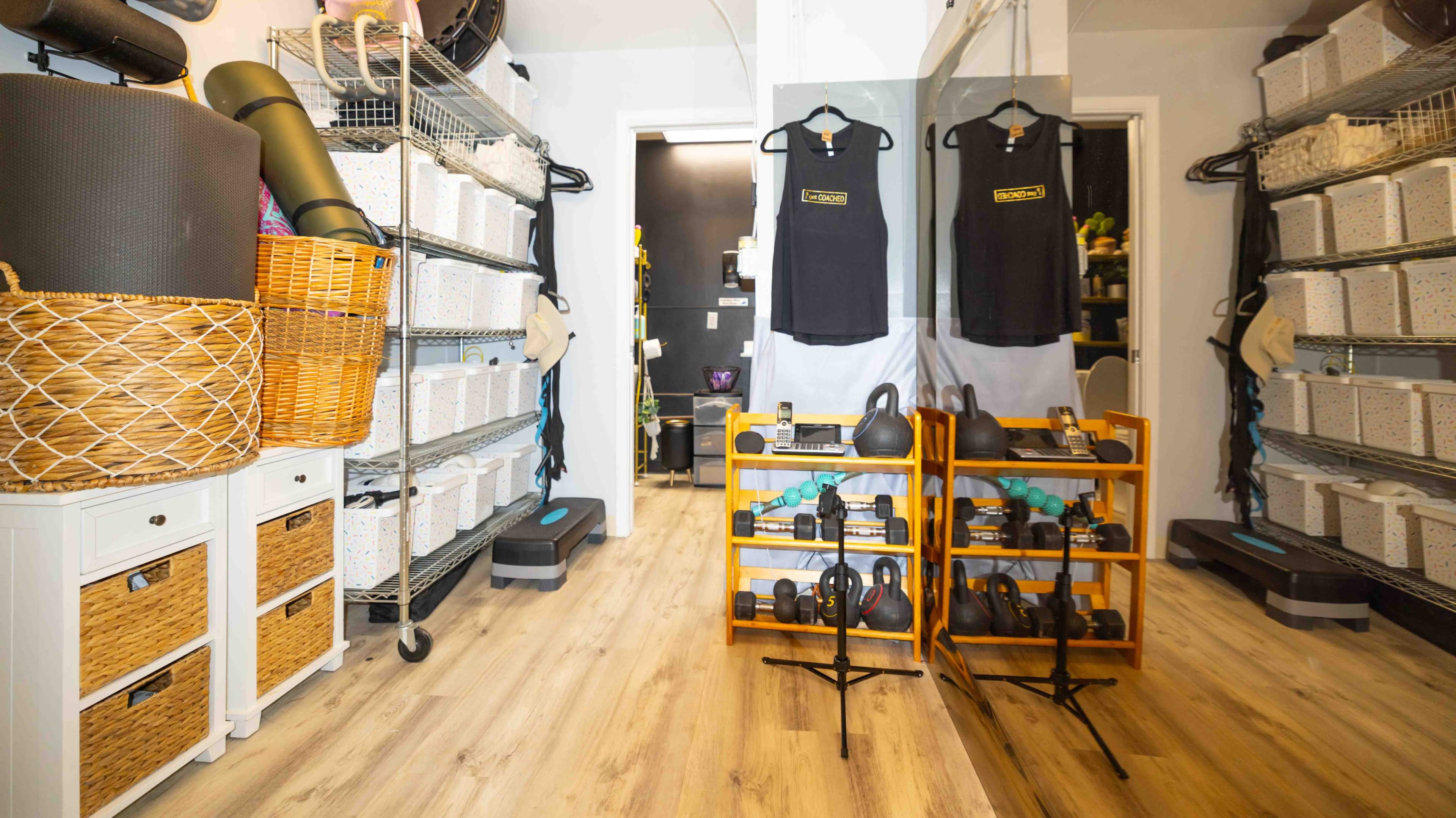 The image shows a home gym area with shelves of storage bins, a mirror, and a wooden rack displaying dumbbells and kettlebells.