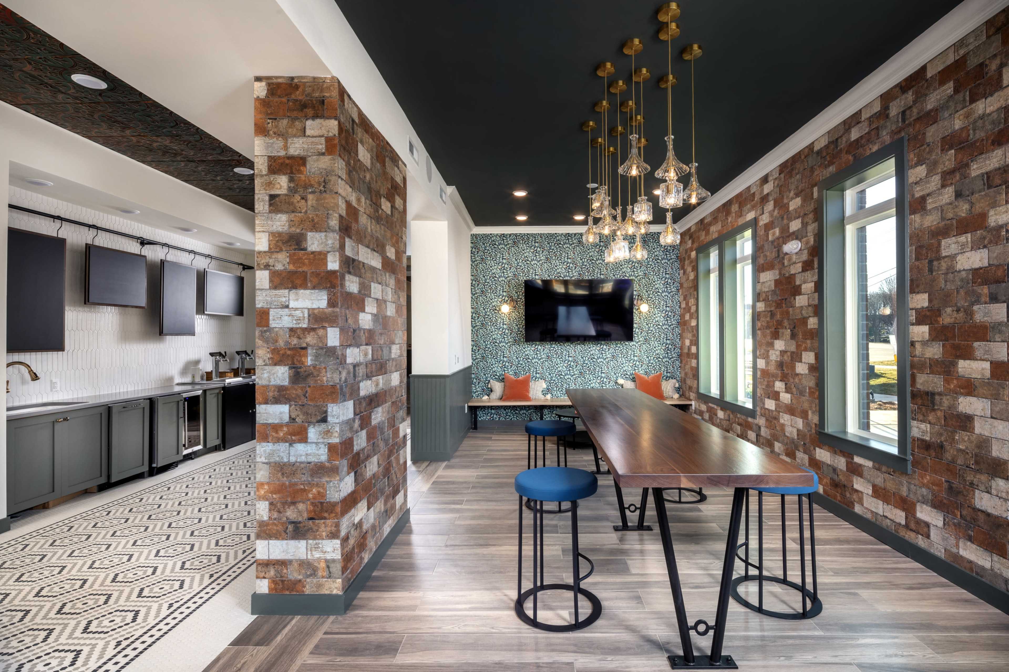A modern communal space featuring a long wooden table with high stools, surrounded by patterned walls, a kitchen area, and decorative lighting fixtures.