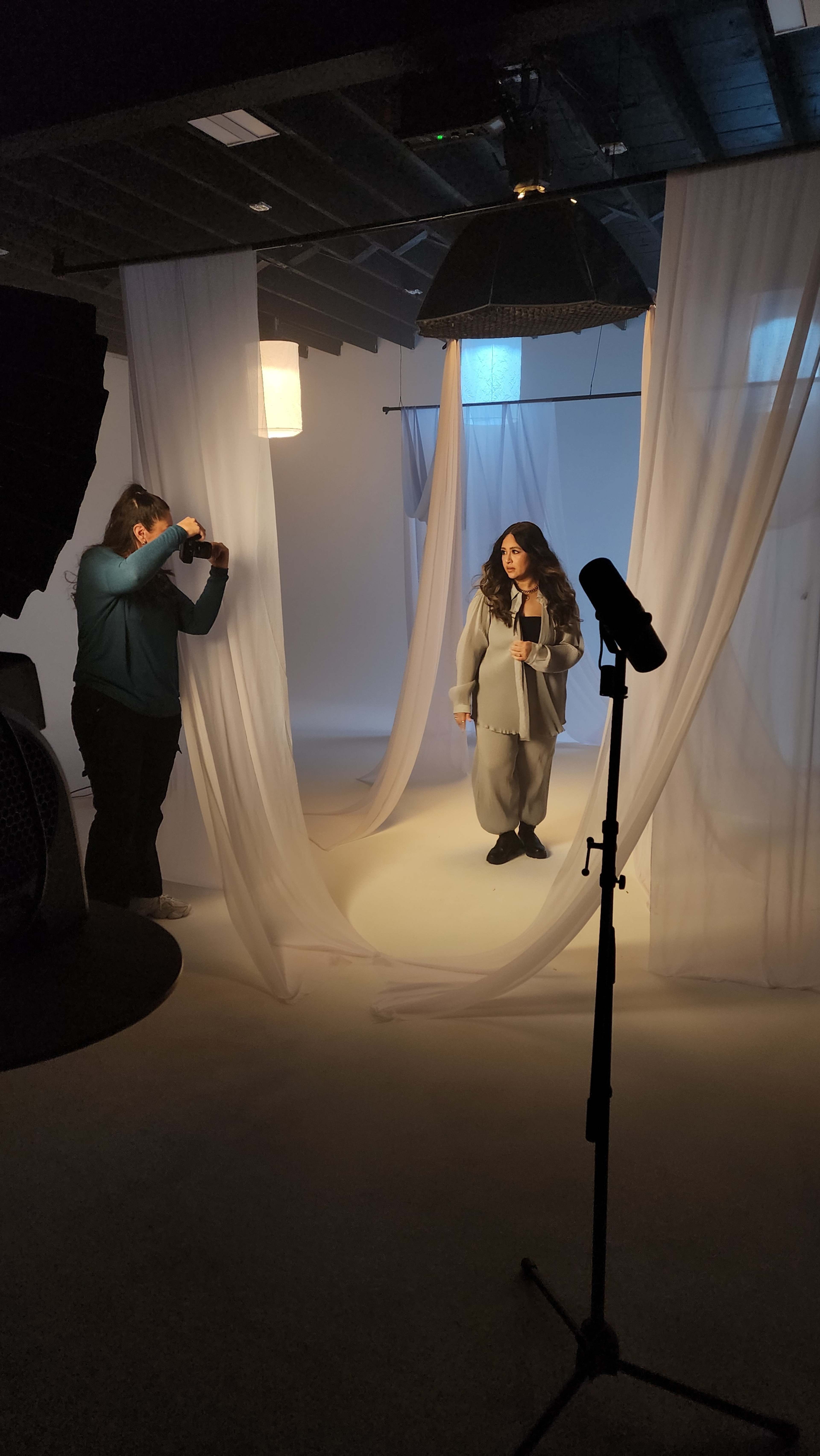 A woman poses in a studio setting surrounded by flowing fabric, while a photographer captures the moment.