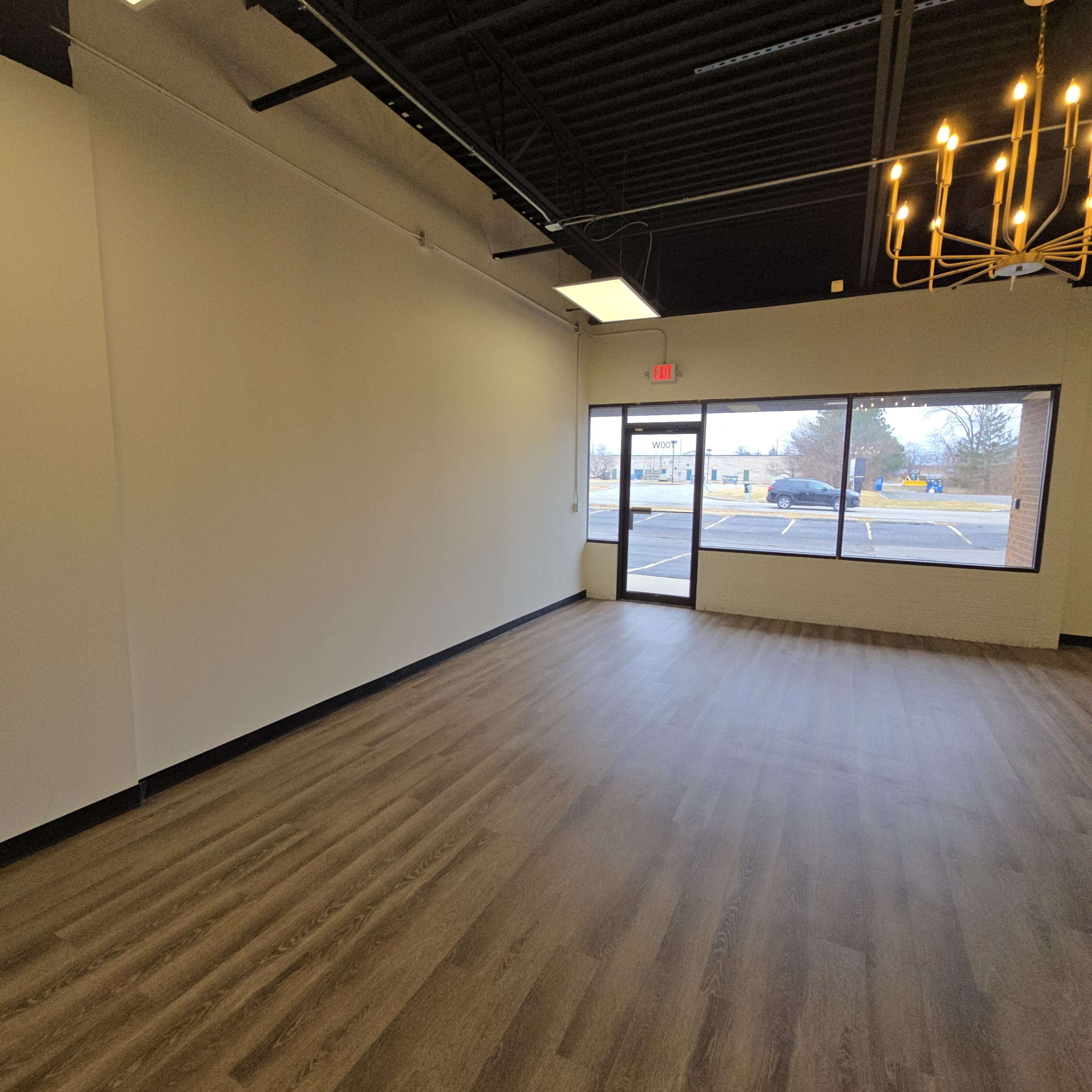 The image shows an empty interior space with light-colored walls, hardwood flooring, and a large window overlooking an outdoor parking area.