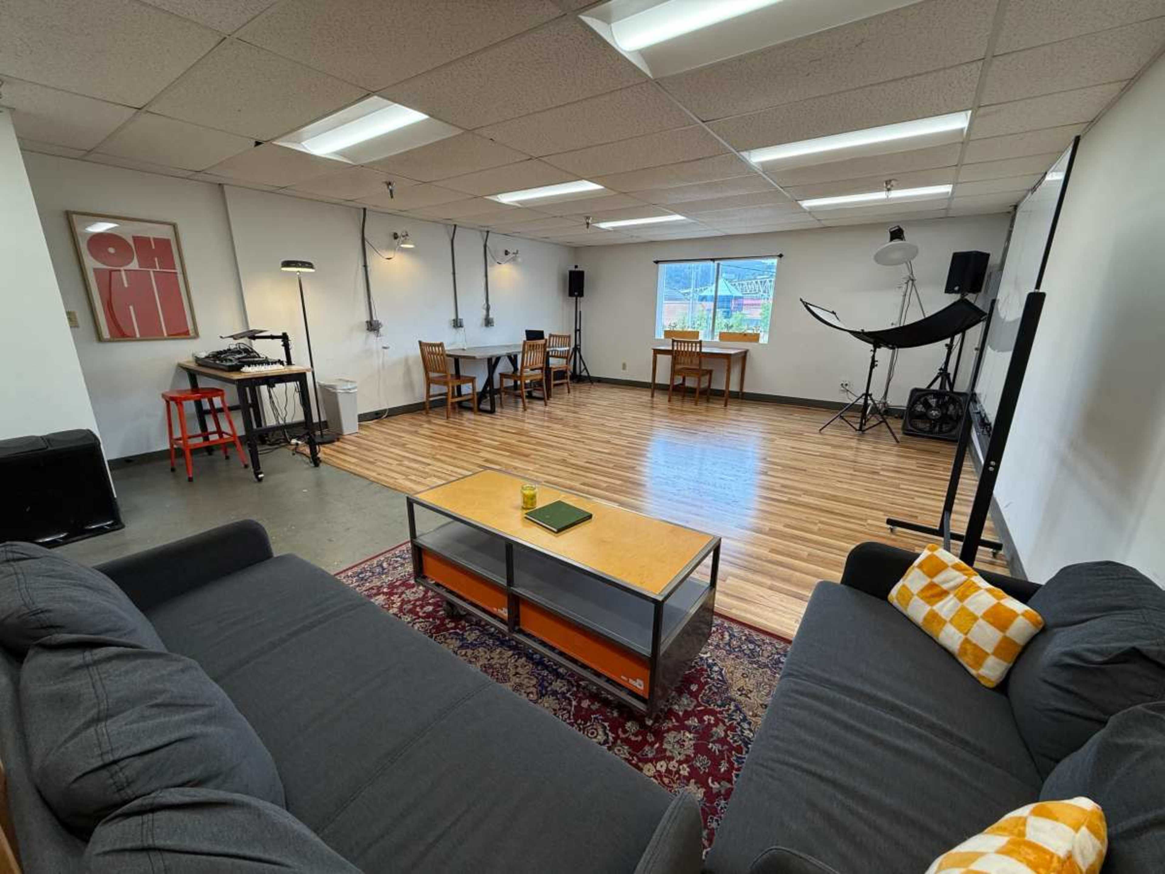 The image shows a spacious room with wooden flooring, featuring two gray couches, a coffee table, a dining table with chairs, and various lighting equipment.