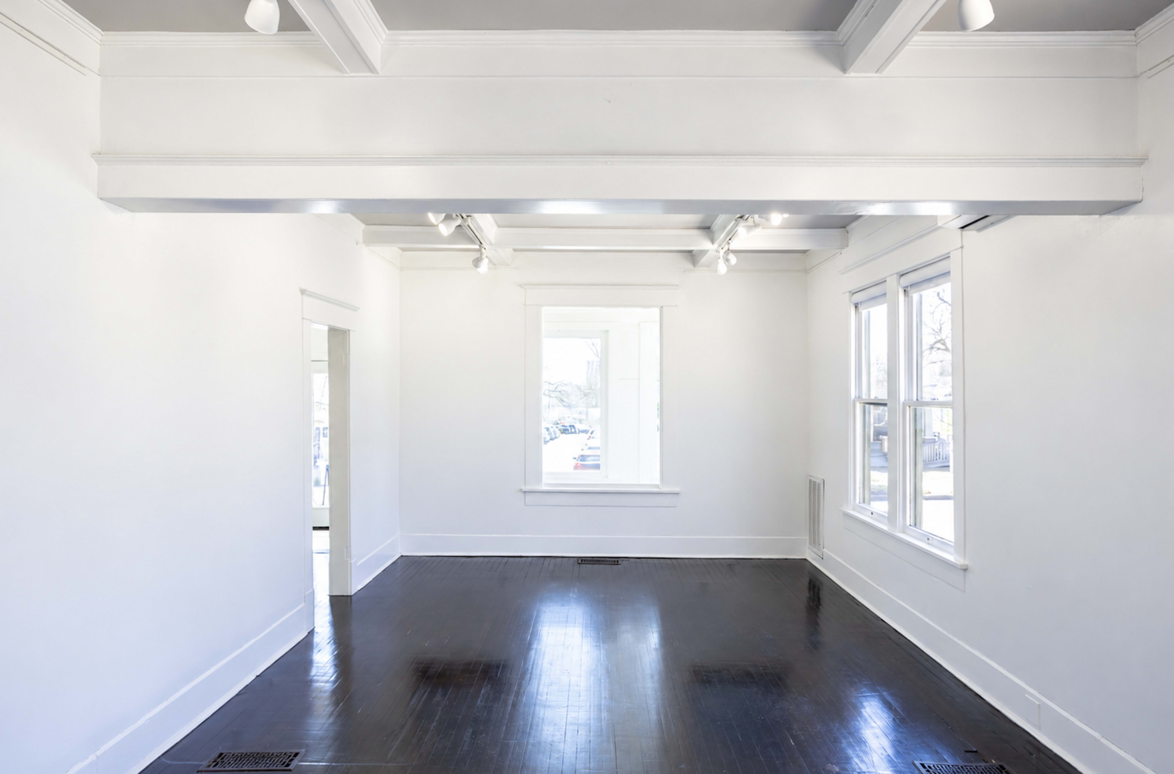 The image shows a spacious, empty room with white walls, a polished dark wooden floor, and natural light streaming in through large windows.