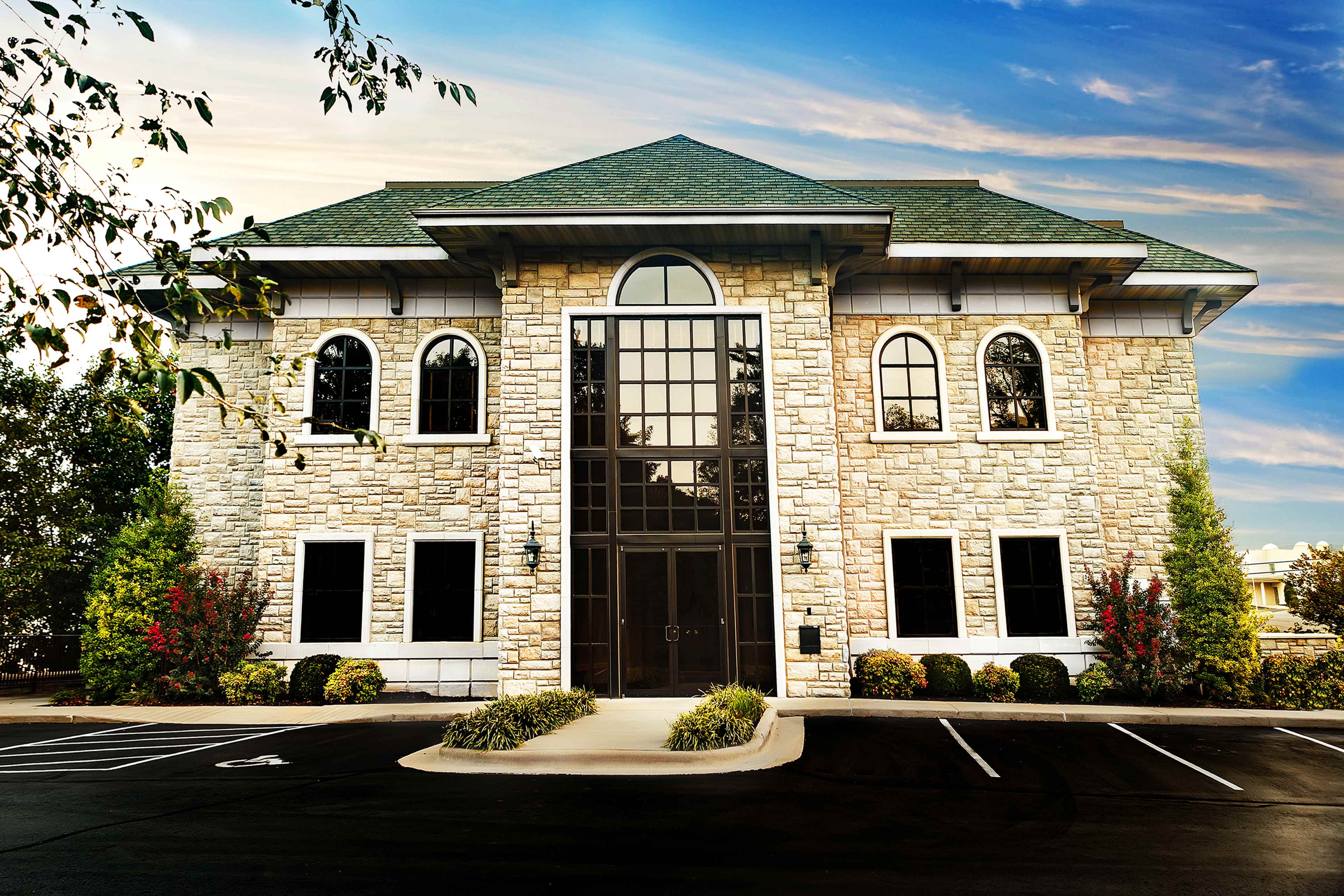 A large stone building with arched windows and a triangular roof is situated in a landscaped parking lot.
