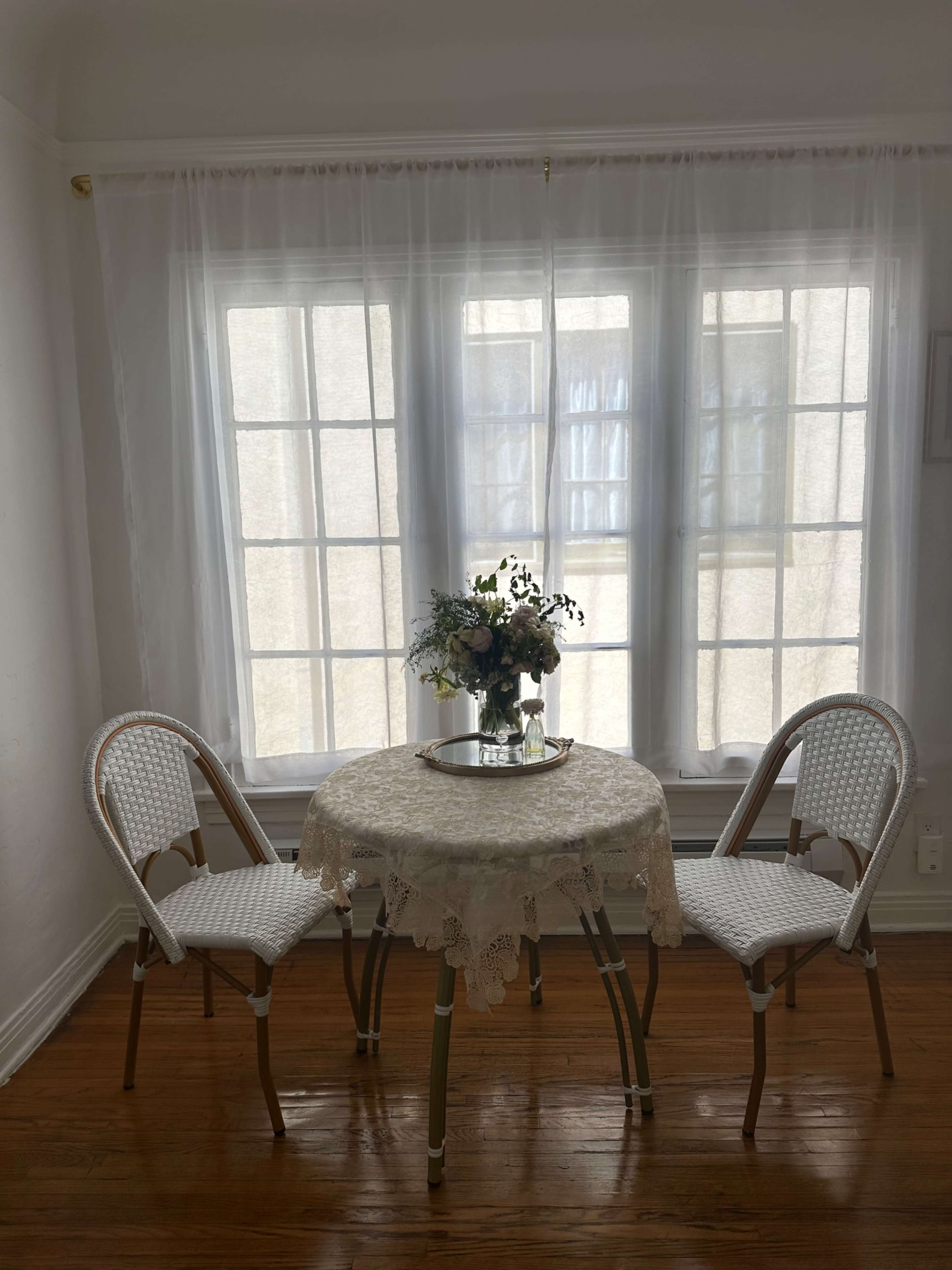 A small round table with a lace tablecloth and a vase of flowers is positioned between two wicker chairs in a sunlit room with large windows.