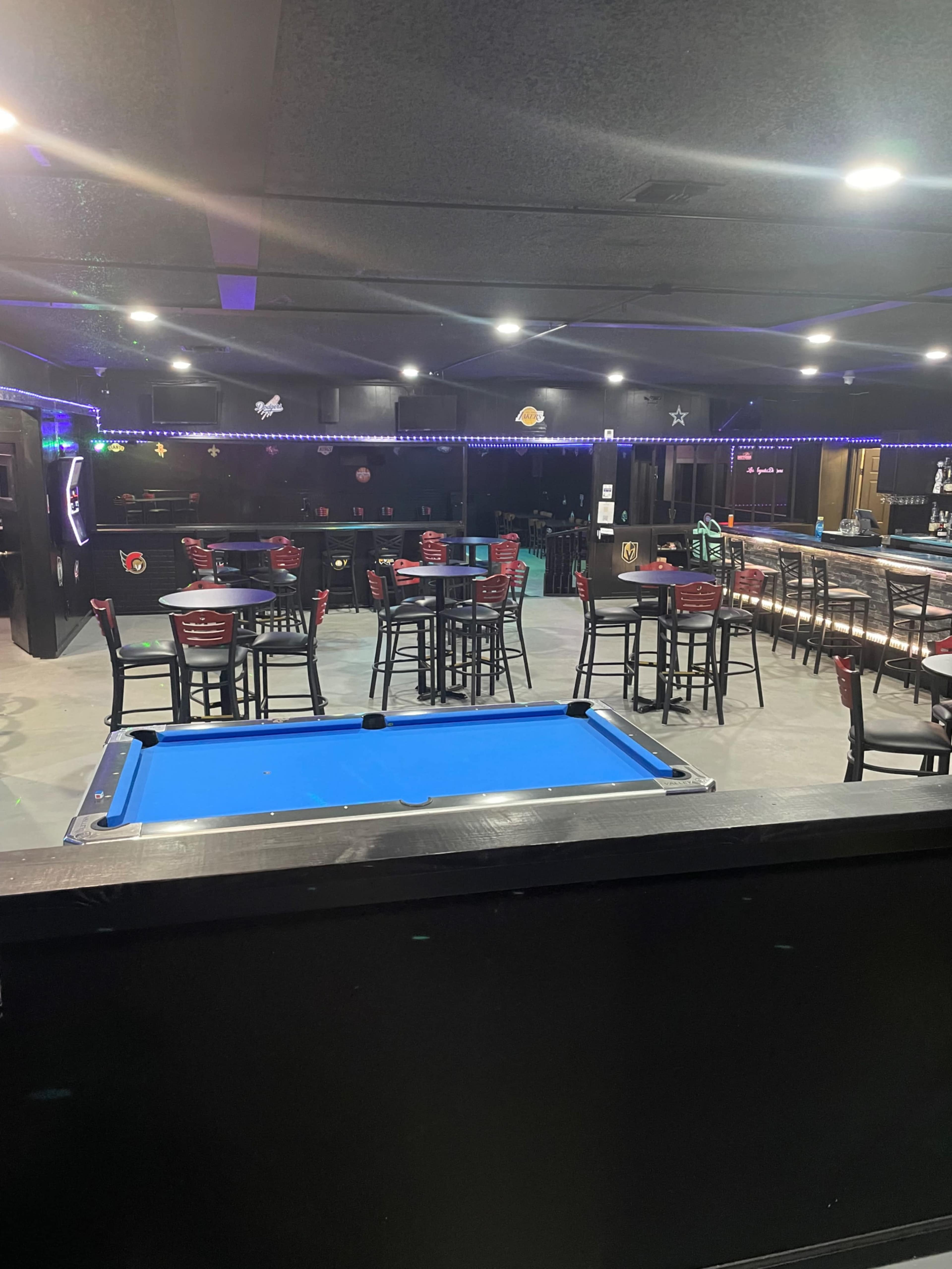 The image shows a dimly lit bar area featuring a blue pool table and several high-top tables with chairs.