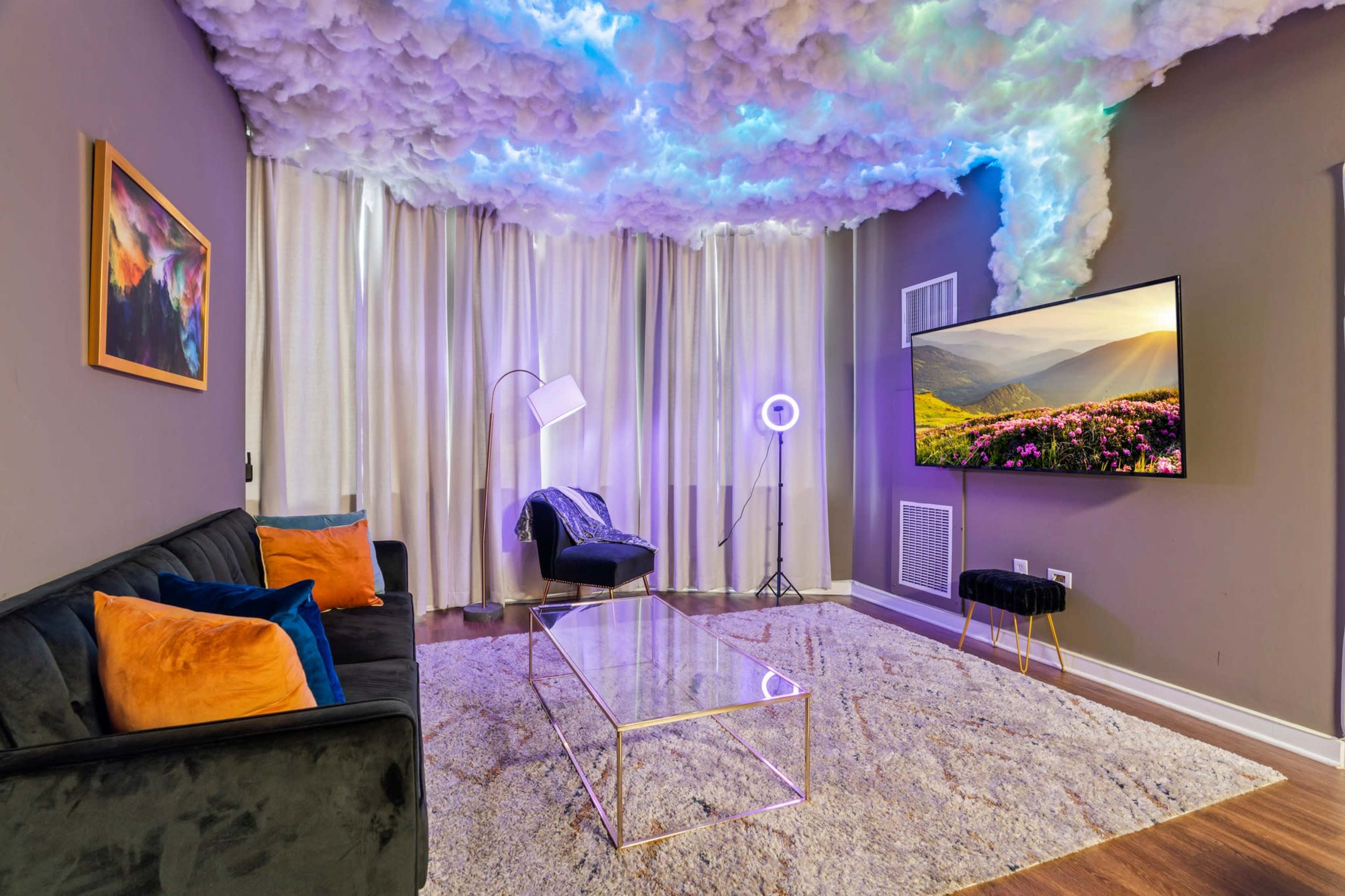 A modern living room features a black couch with colorful pillows, a glass coffee table, a wall-mounted TV, and a ceiling designed to resemble clouds with blue lighting.