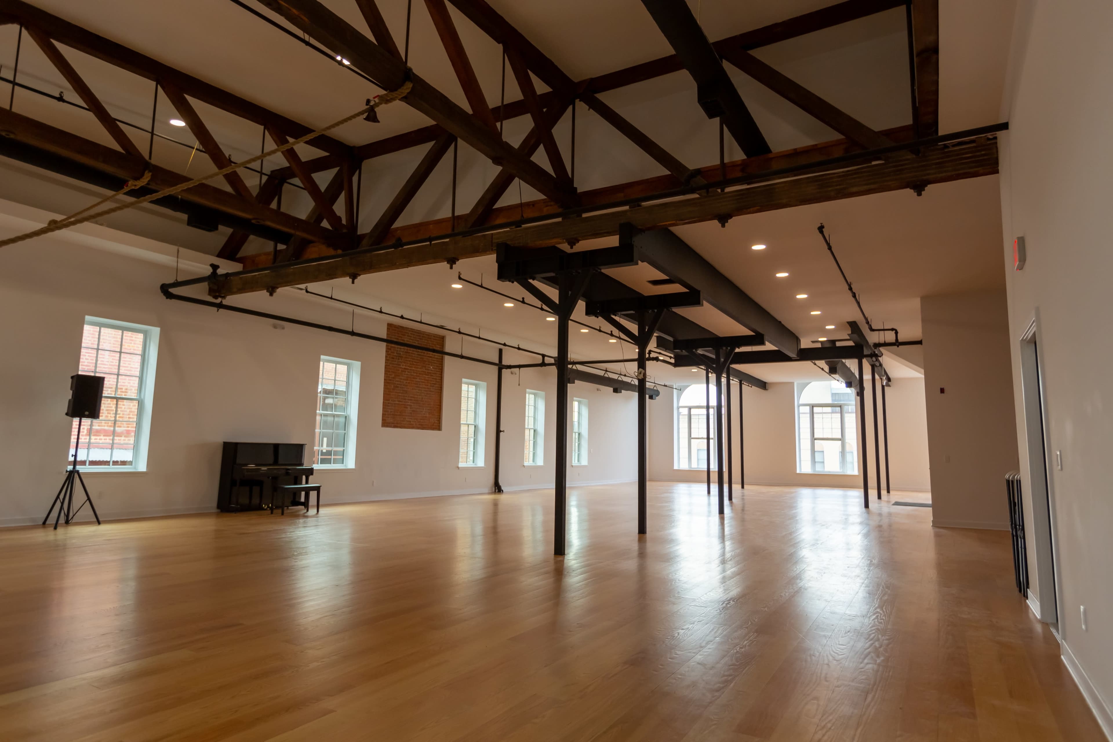 A spacious empty room with wooden flooring, exposed beams, and large windows, featuring a piano in one corner.