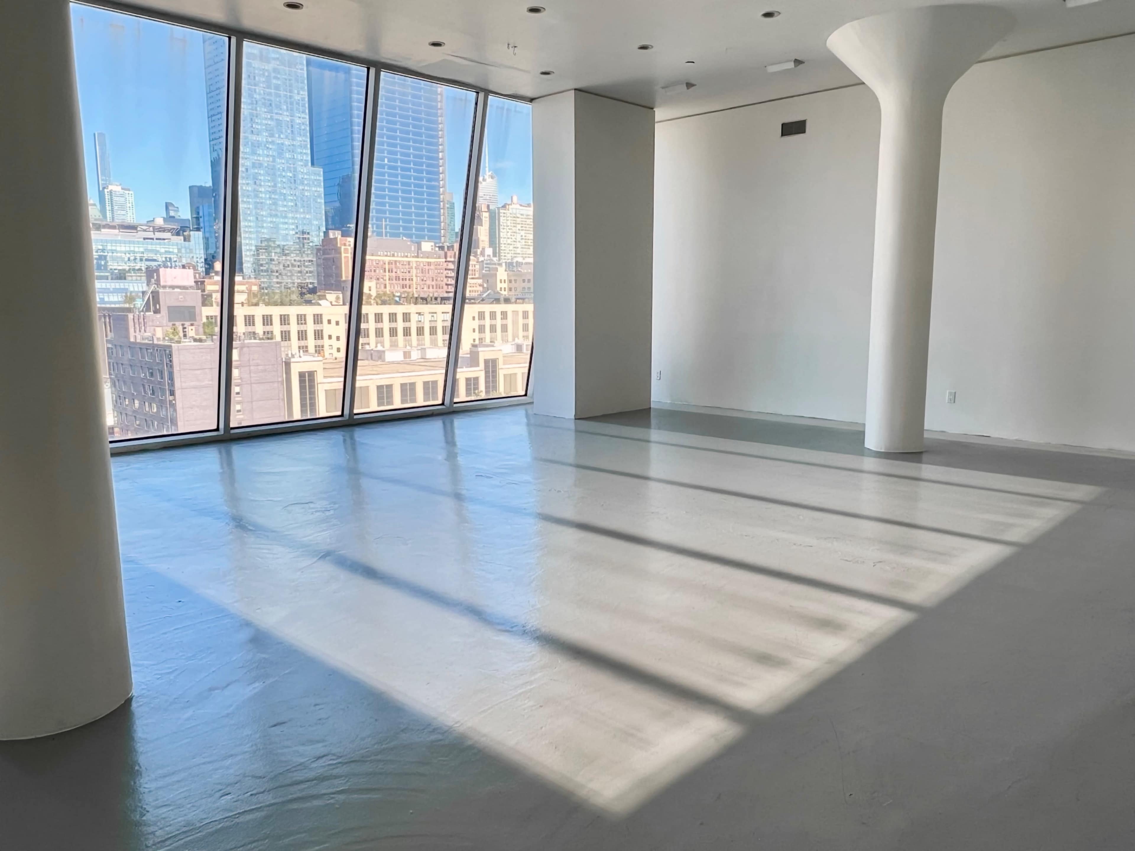 A spacious room with large windows providing a view of city buildings and a concrete floor illuminated by natural light.