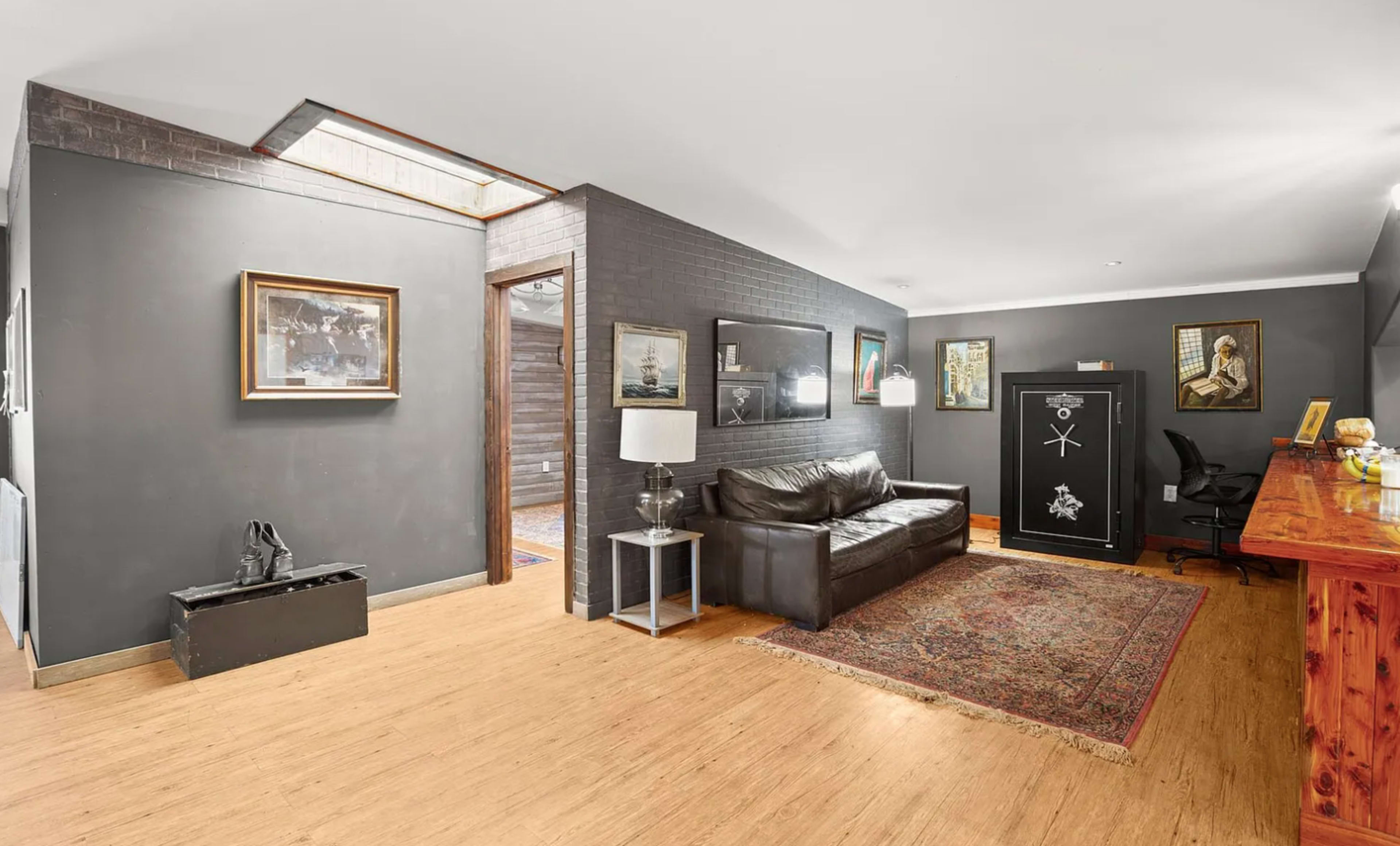 The image shows a modern living area with a dark wall, a leather sofa, artwork, a wooden console table, and a large safe.