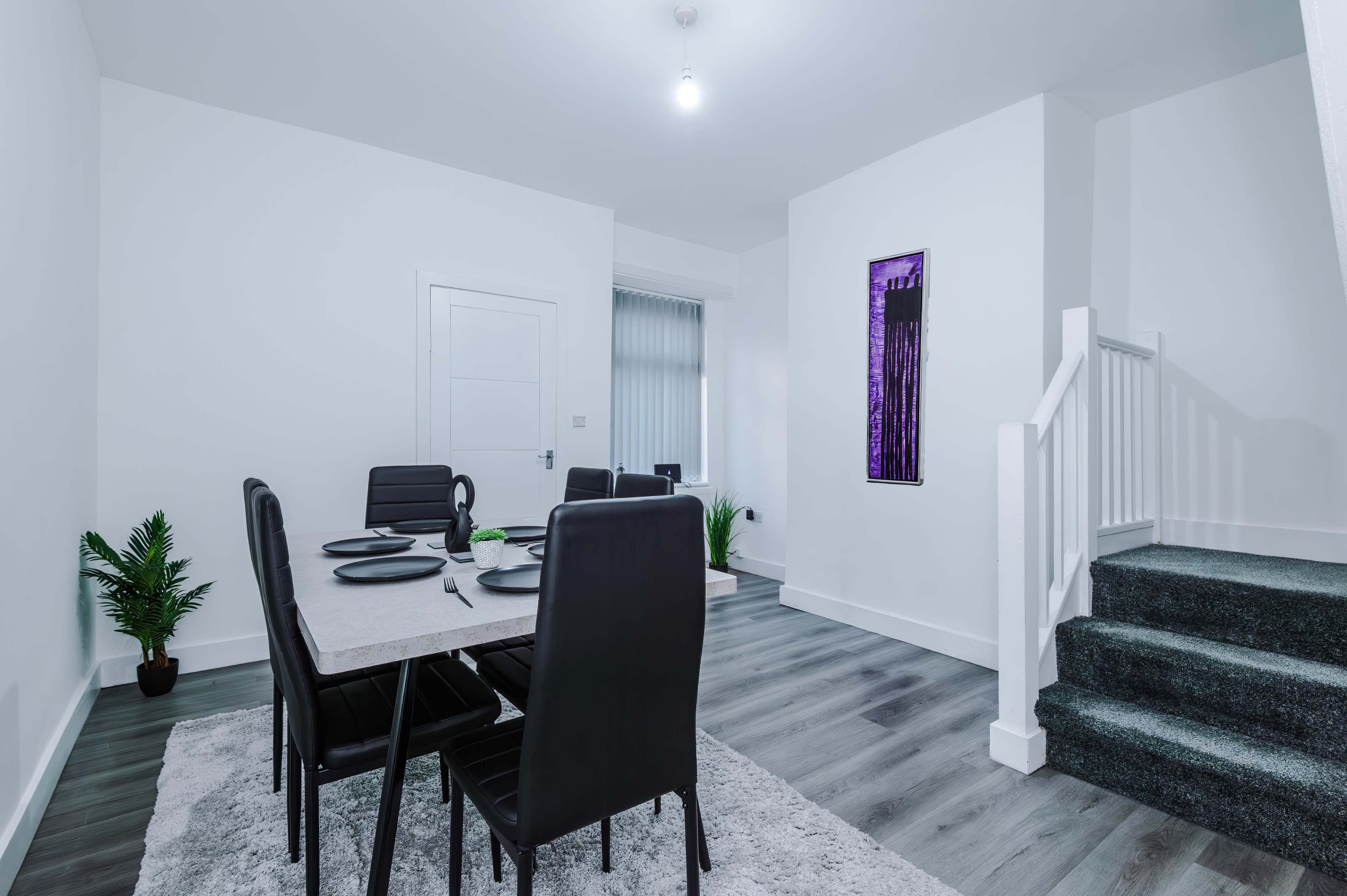 A dining area features a rectangular table set for six, accompanied by a staircase and a piece of contemporary artwork on the wall.