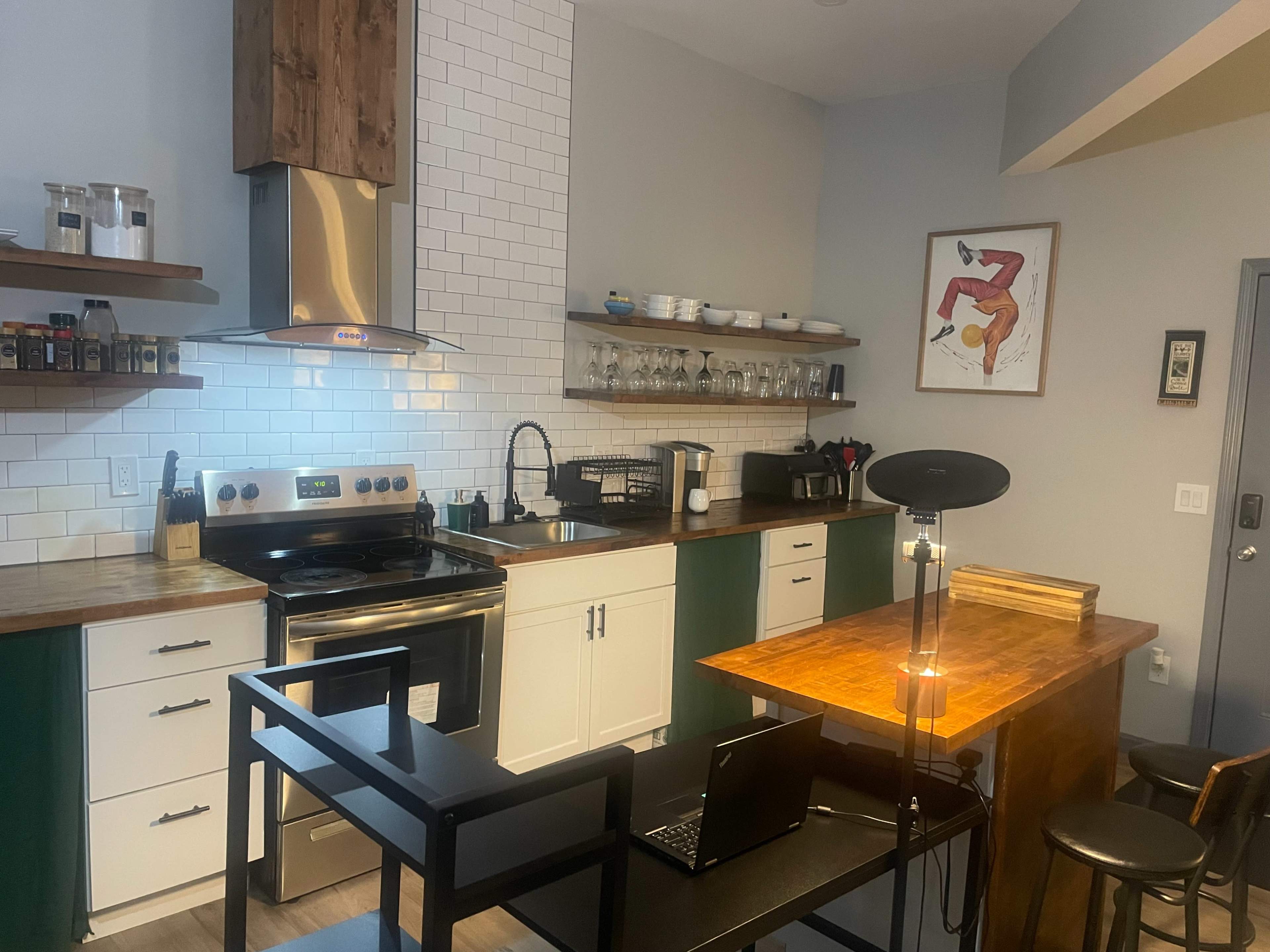 The image shows a modern kitchen with a stainless steel stove, white cabinets, and a wooden countertop, along with a small dining area featuring two stools and a laptop on the table.