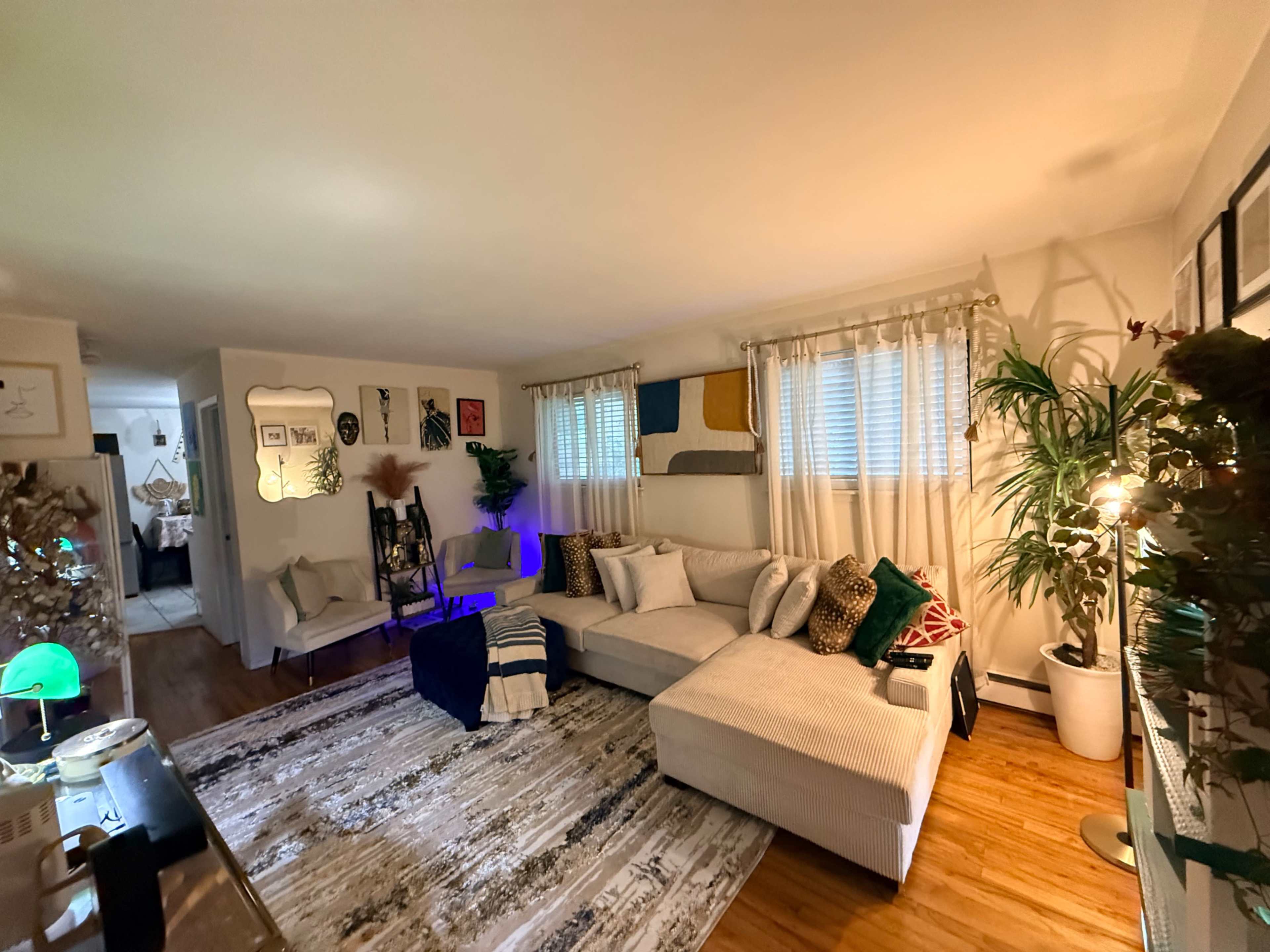 The image shows a cozy living room with a large sectional sofa, a patterned area rug, and decorative pillows, illuminated by natural light through window curtains.