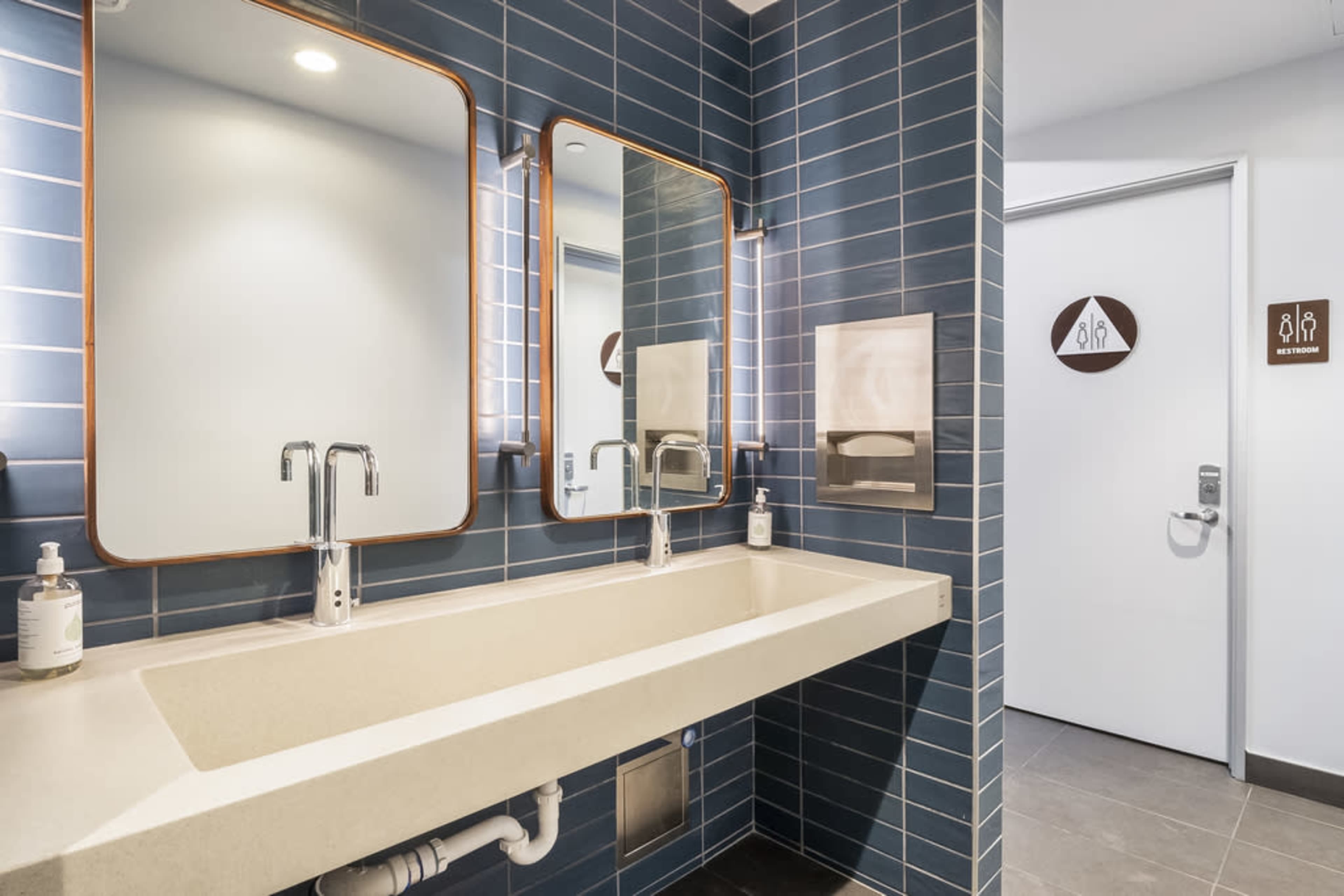The image shows a modern restroom with a long, minimalist sink, two mirrors, and a door leading to the restroom area.