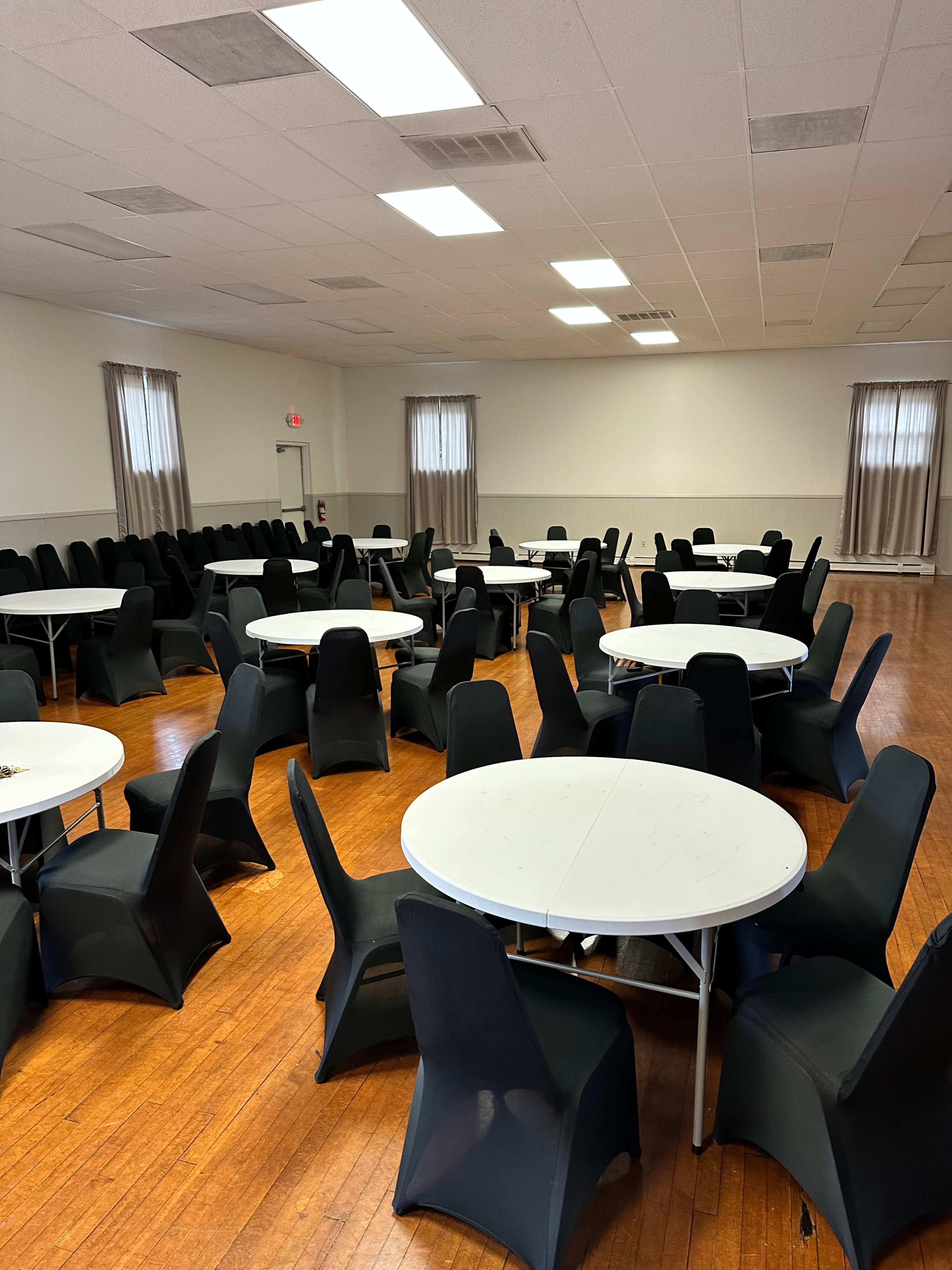 A spacious hall contains several round tables surrounded by black chairs on a polished wooden floor.