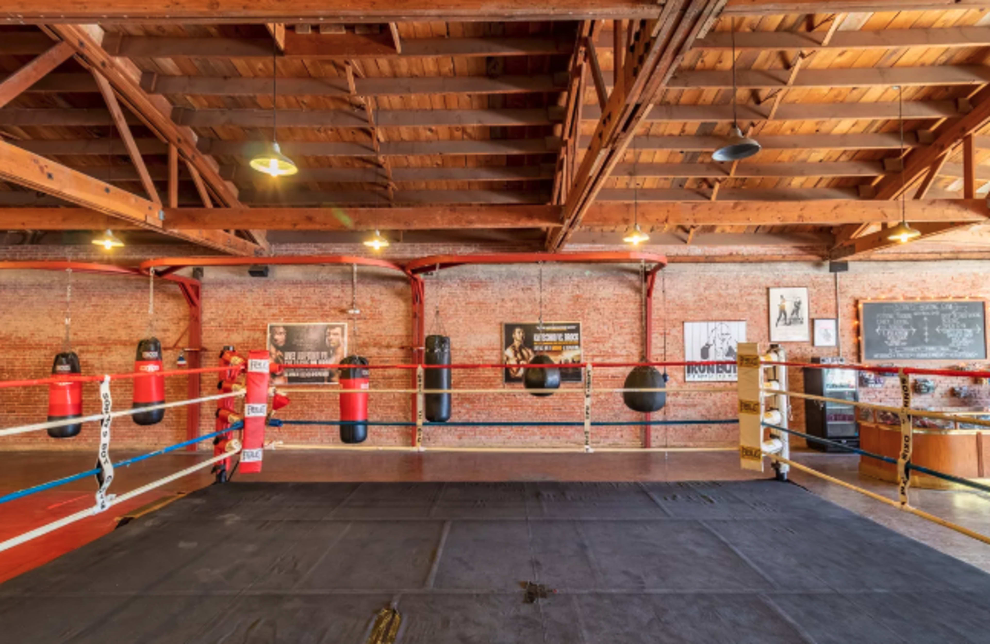 The image shows a boxing gym featuring a ring surrounded by heavy bags and adorned with vintage boxing posters on the brick walls.