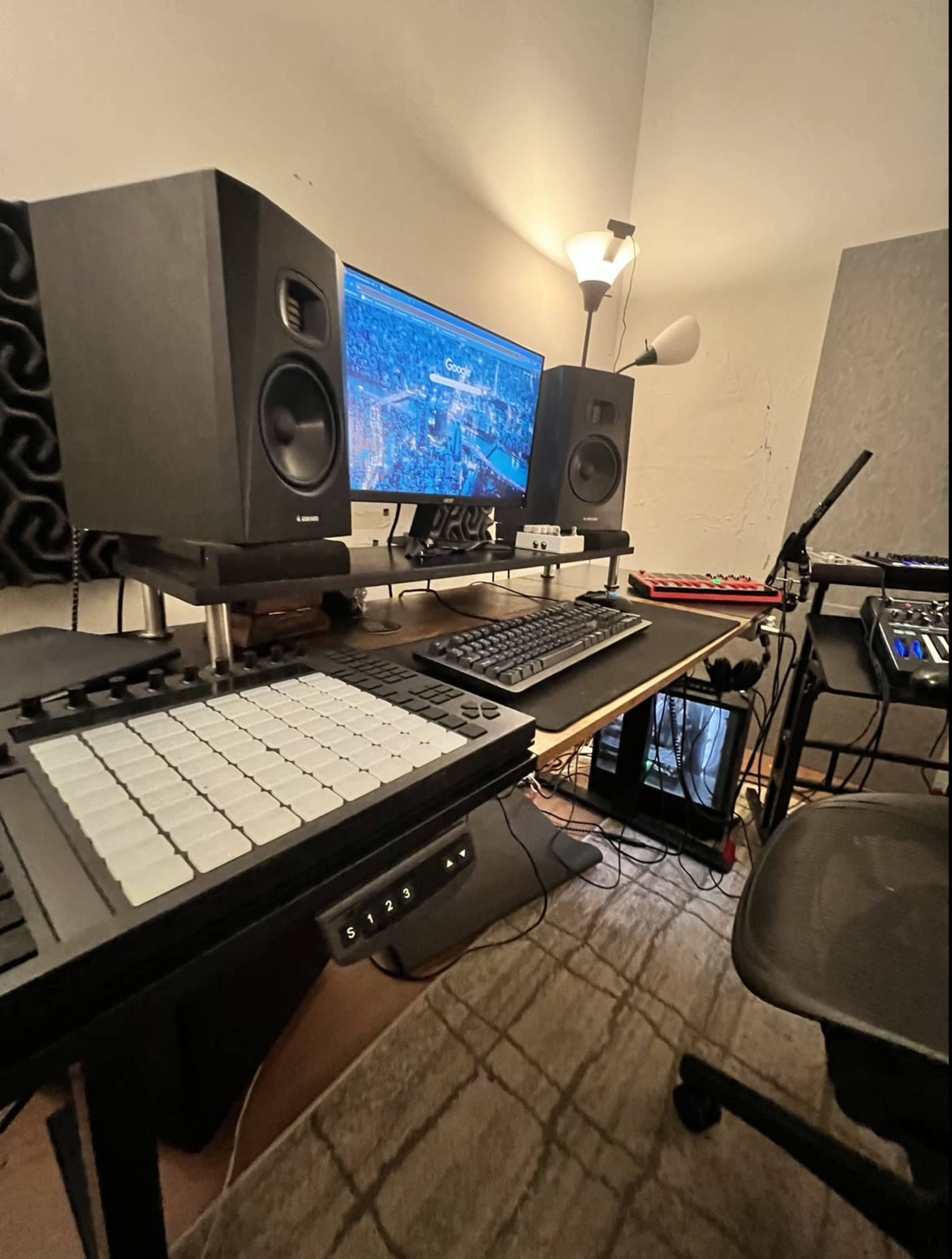 A music production setup featuring a computer monitor, audio speakers, a MIDI controller, and a keyboard on a desk in a well-lit room.