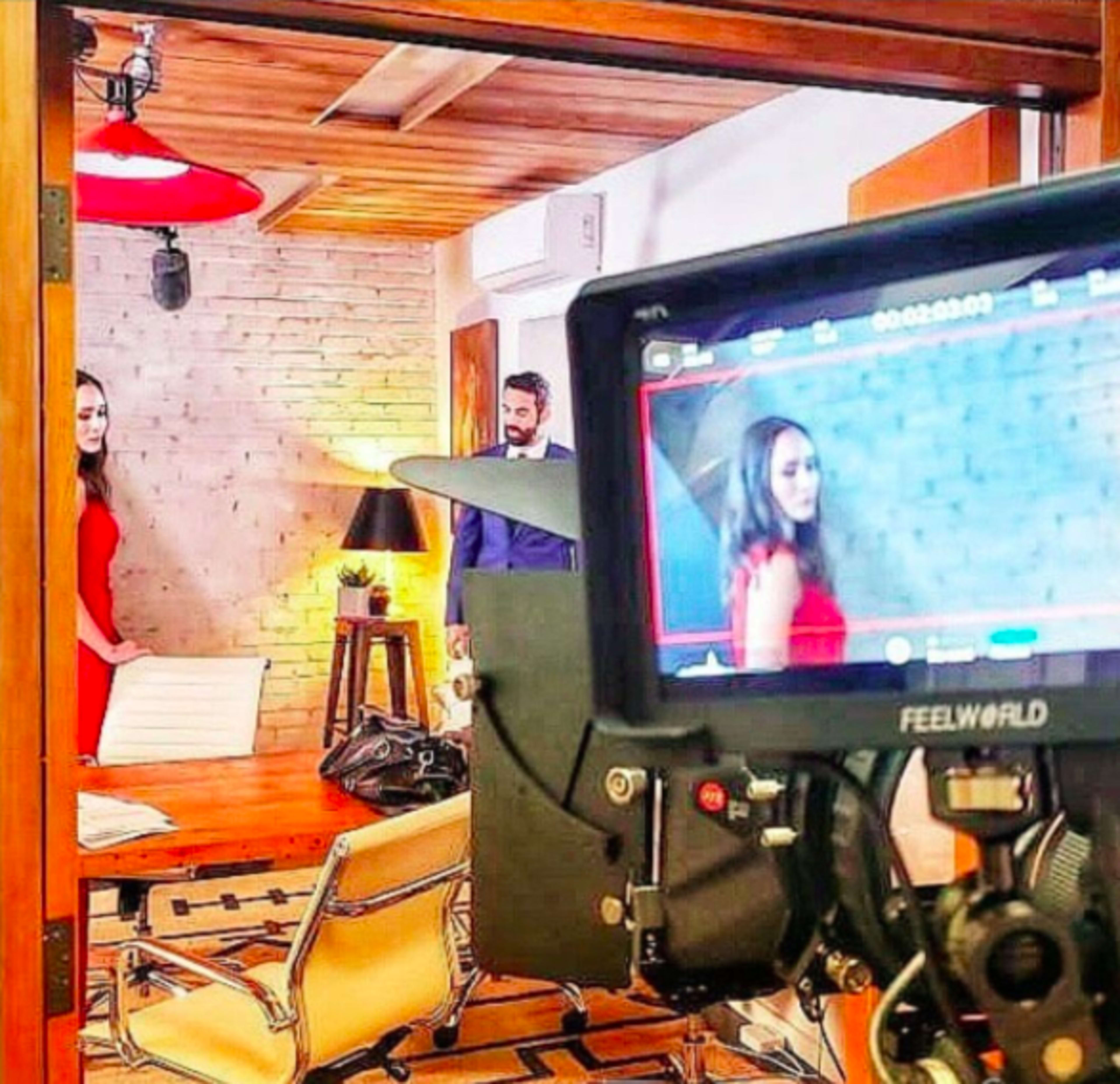 A film crew is positioned in an indoor setting, capturing a scene with two actors, one in a red dress and another in formal attire, in front of a brick wall.