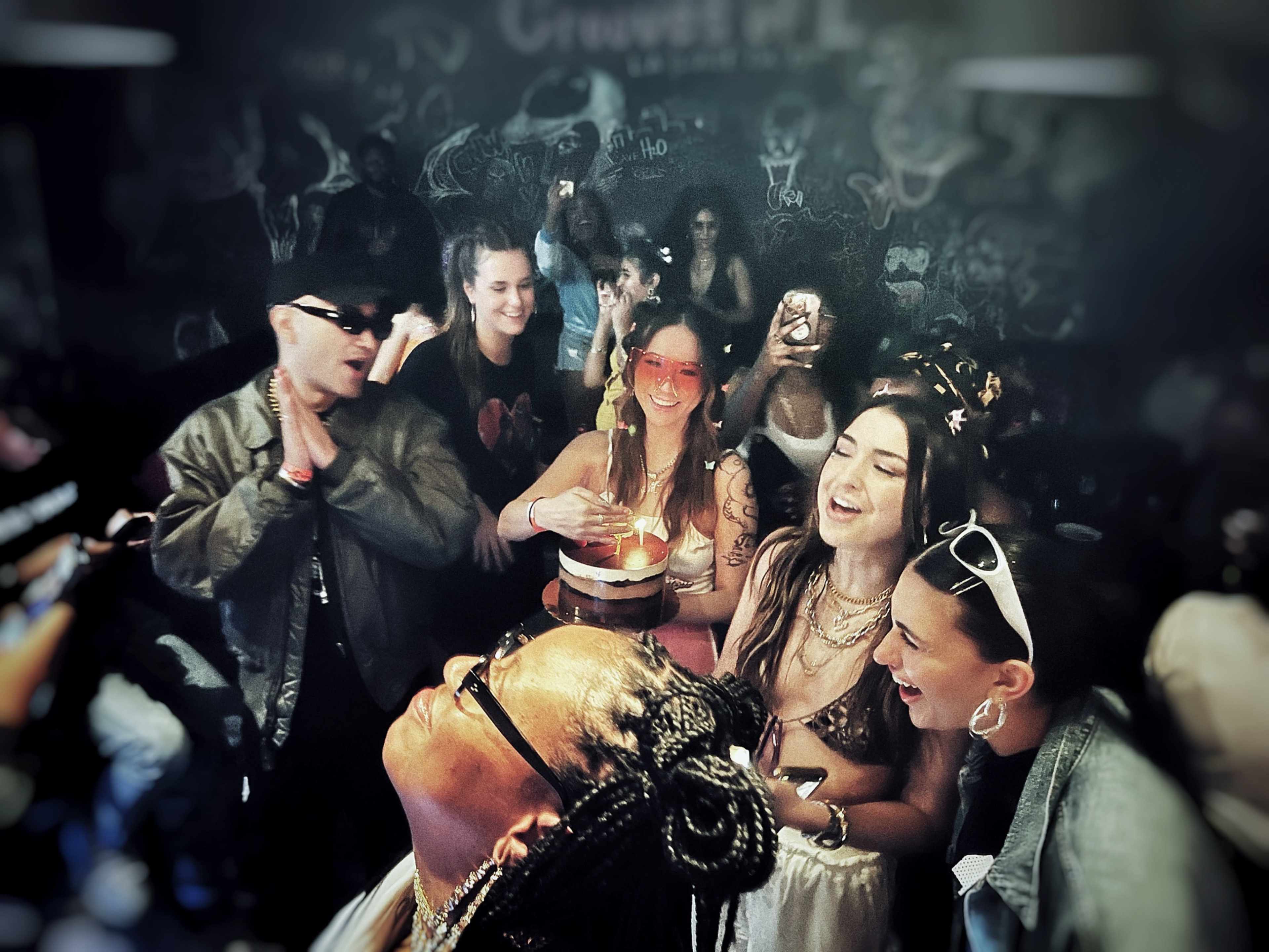 A group of people celebrate in a dimly lit venue, surrounded by dark graffiti-covered walls, with some individuals holding drinks and a cake.