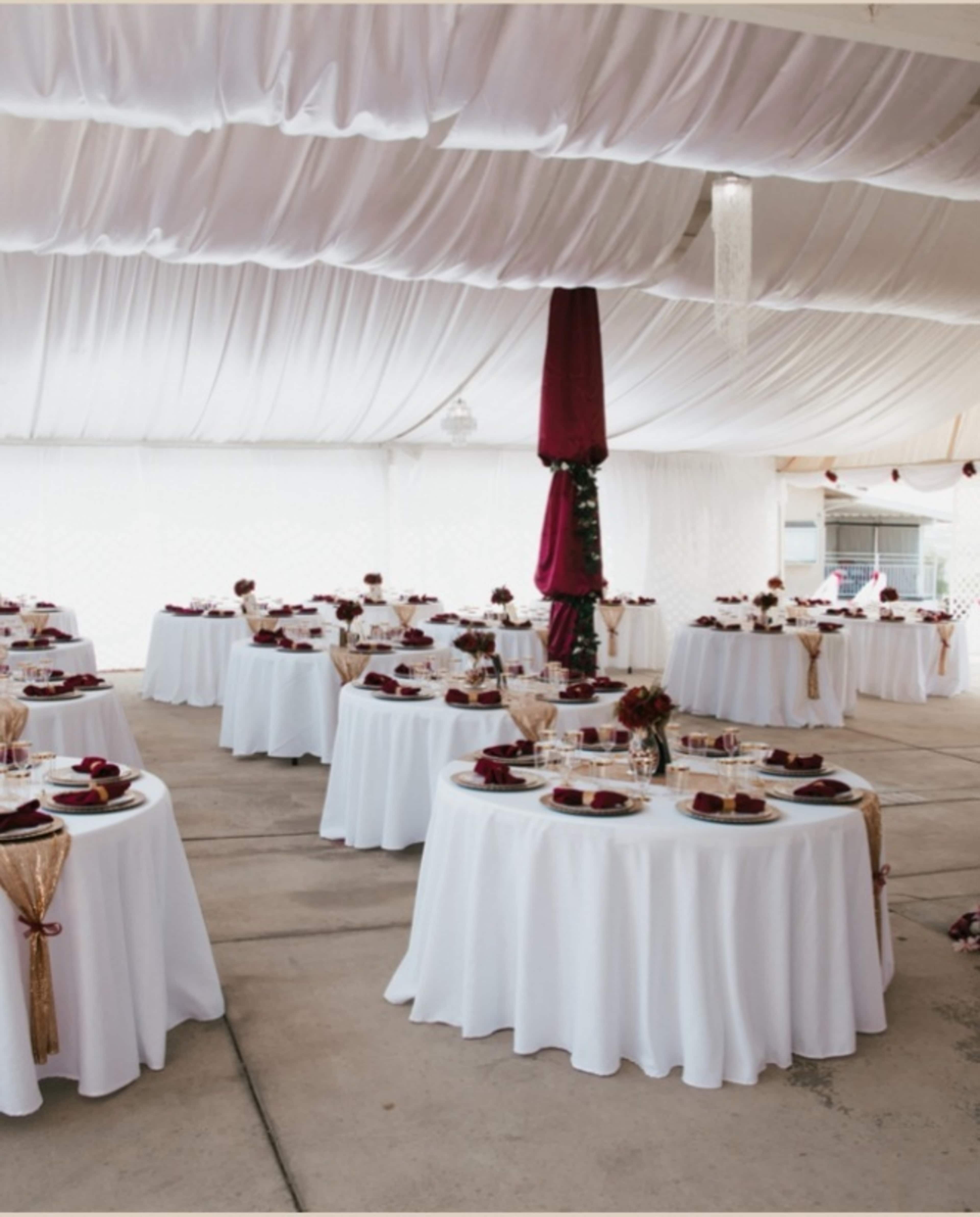 A large tent is set up with round tables covered in white tablecloths, each adorned with plates, glasses, and floral centerpieces.
