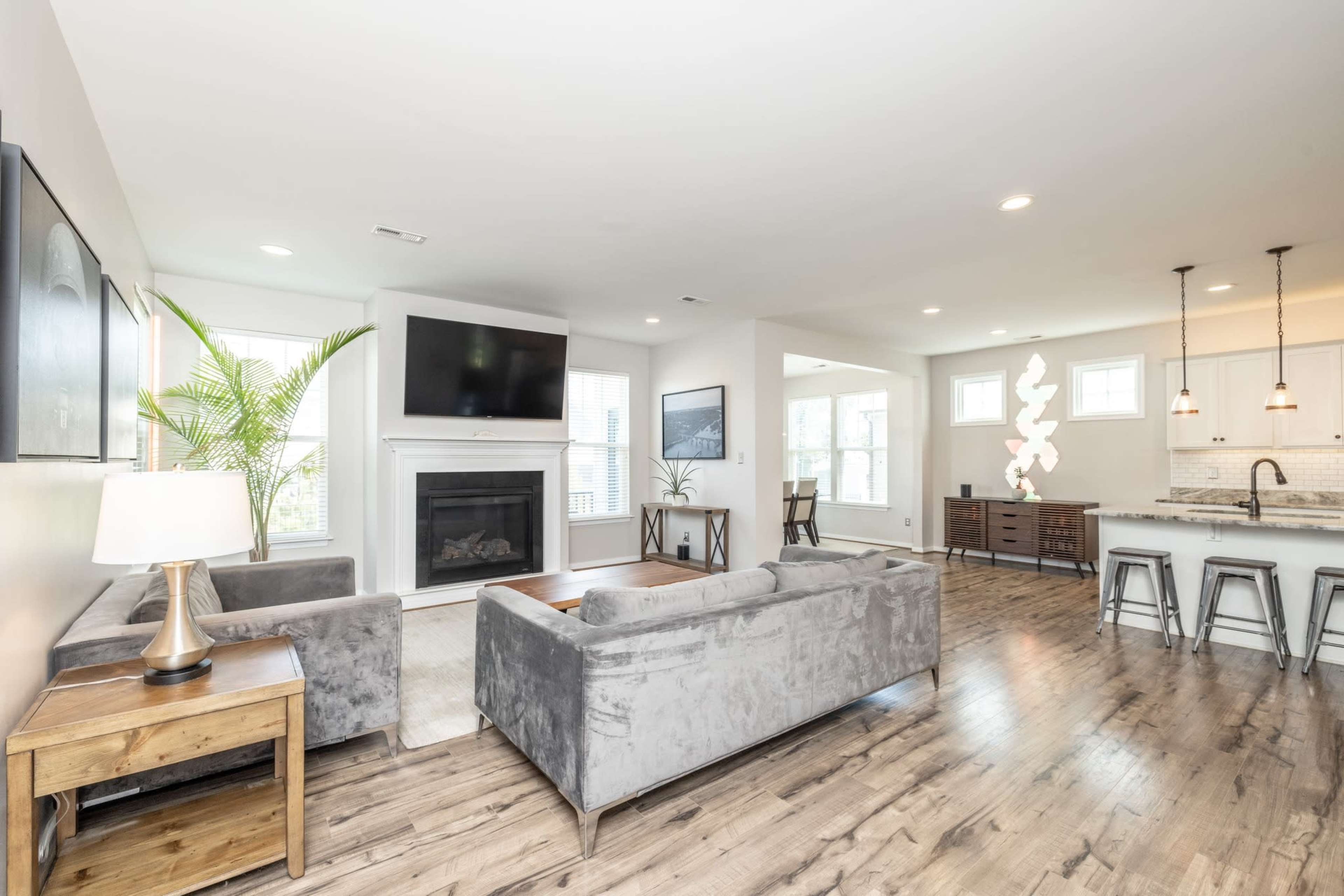 A modern living room with a fireplace, a large television mounted on the wall, and an open kitchen featuring sleek bar stools and a dining area.