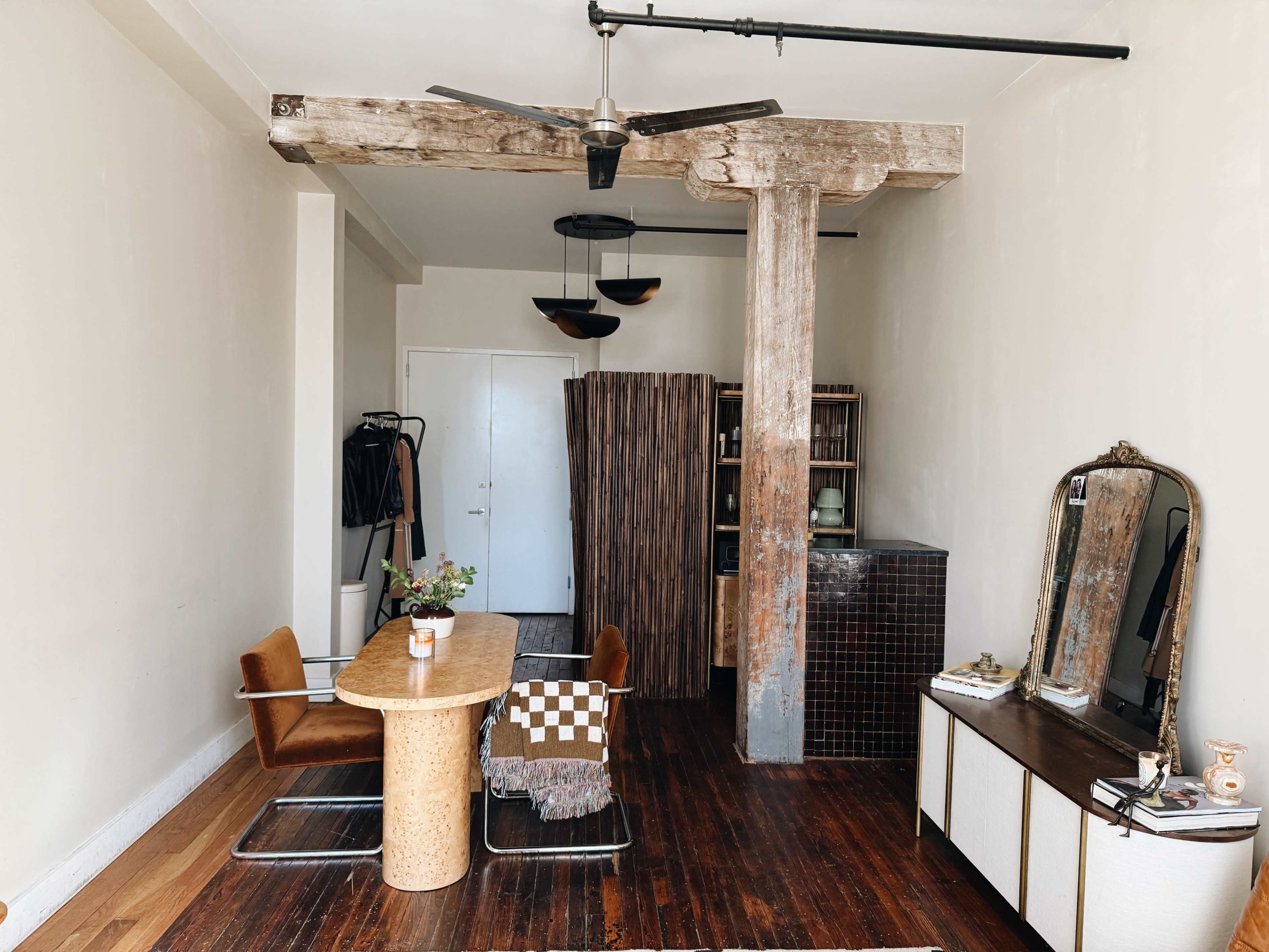 A minimalist living space features a wooden dining table with a patterned cloth, surrounded by chairs, and a tall wooden column, with a mirror and shelves against a wall.