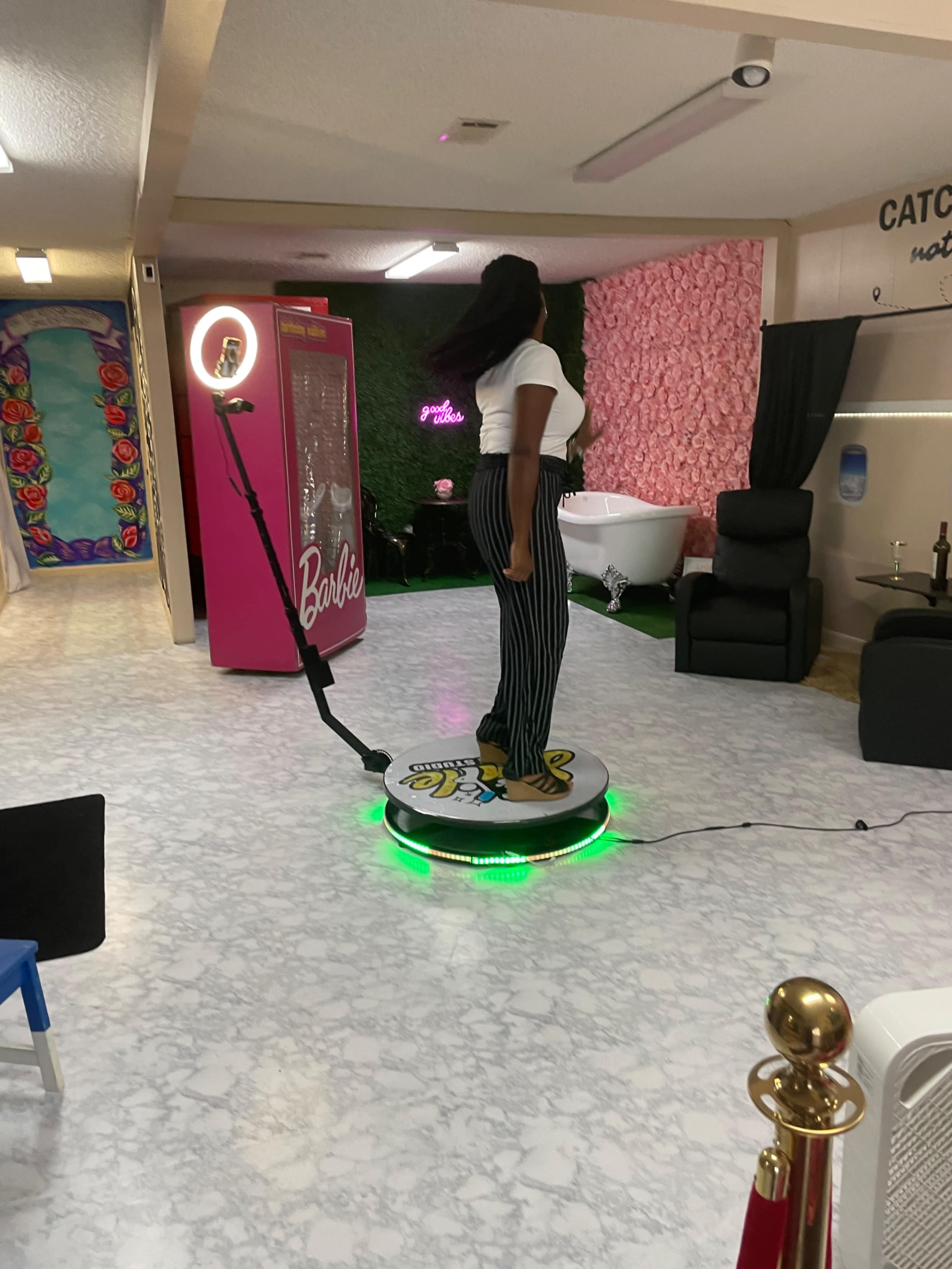 A person stands on a circular platform with lights, surrounded by colorful decor and a vintage-style Barbie-themed display.