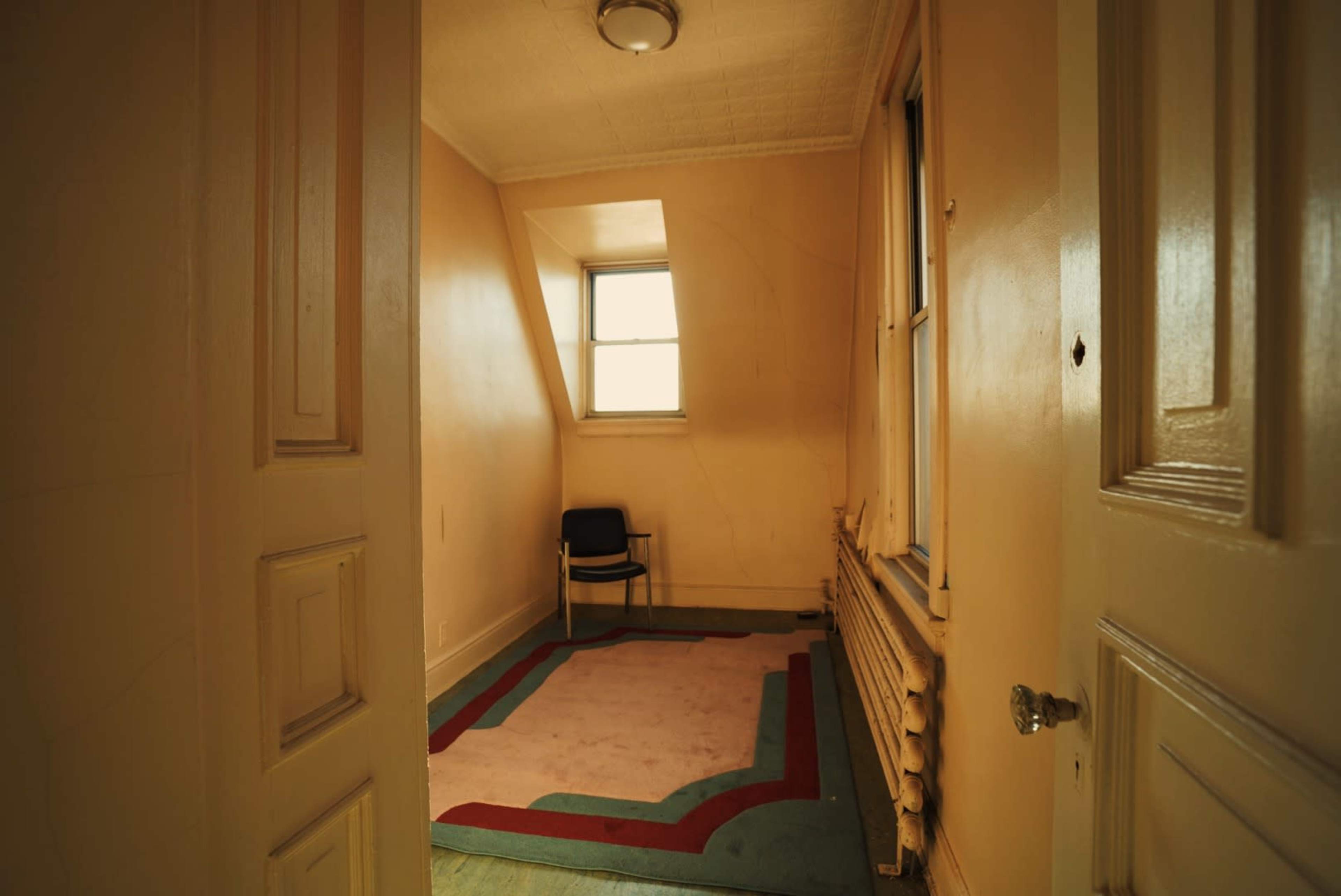 A small, empty room with a single chair, pale yellow walls, and a patterned carpet.