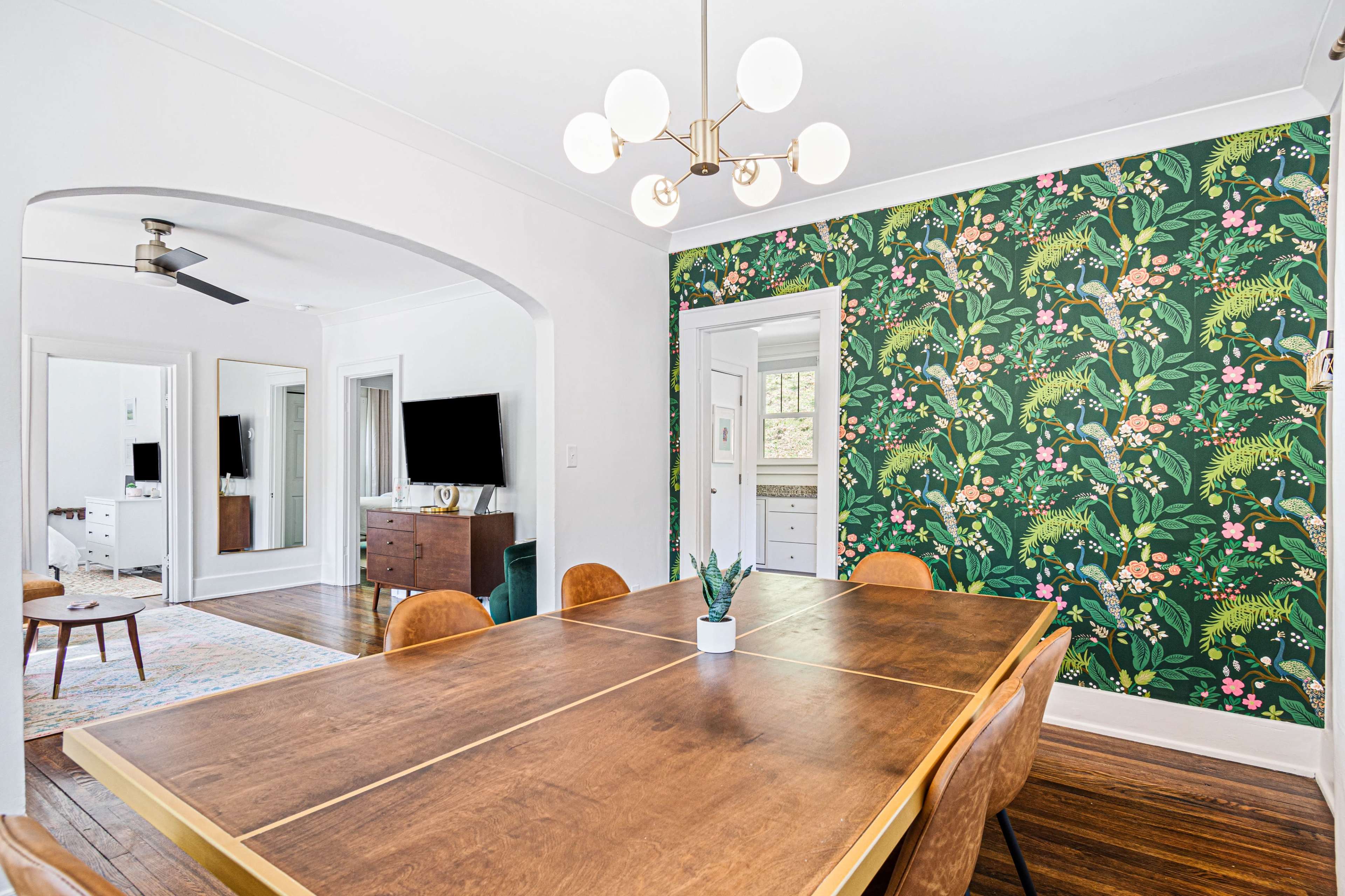 A dining area with a wooden table surrounded by brown chairs, a floral wallpaper on one wall, and a light fixture above the table.