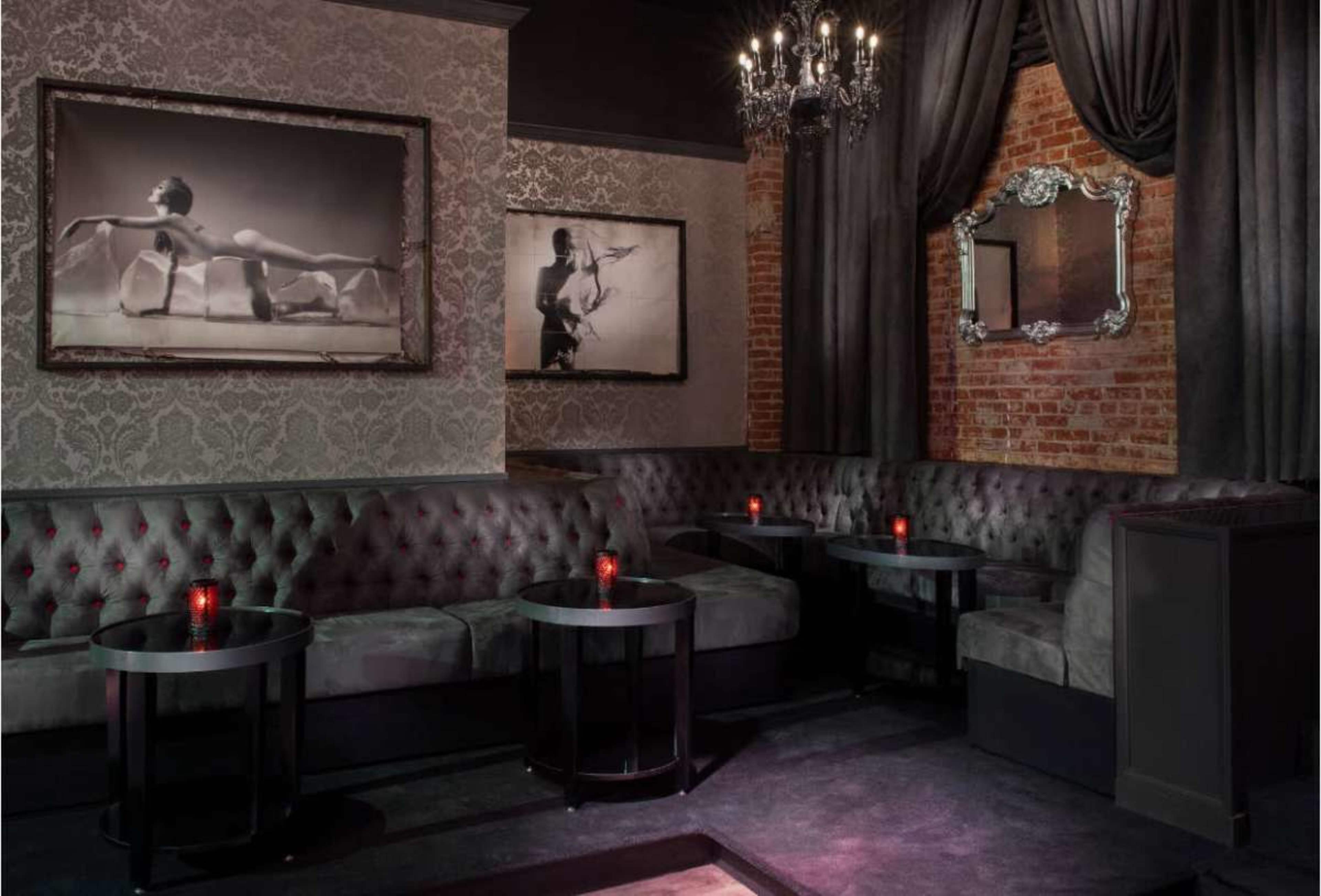 The image shows a stylish lounge area with velvet seating, dim lighting, and black tables, adorned with framed black-and-white photographs and a chandelier overhead.