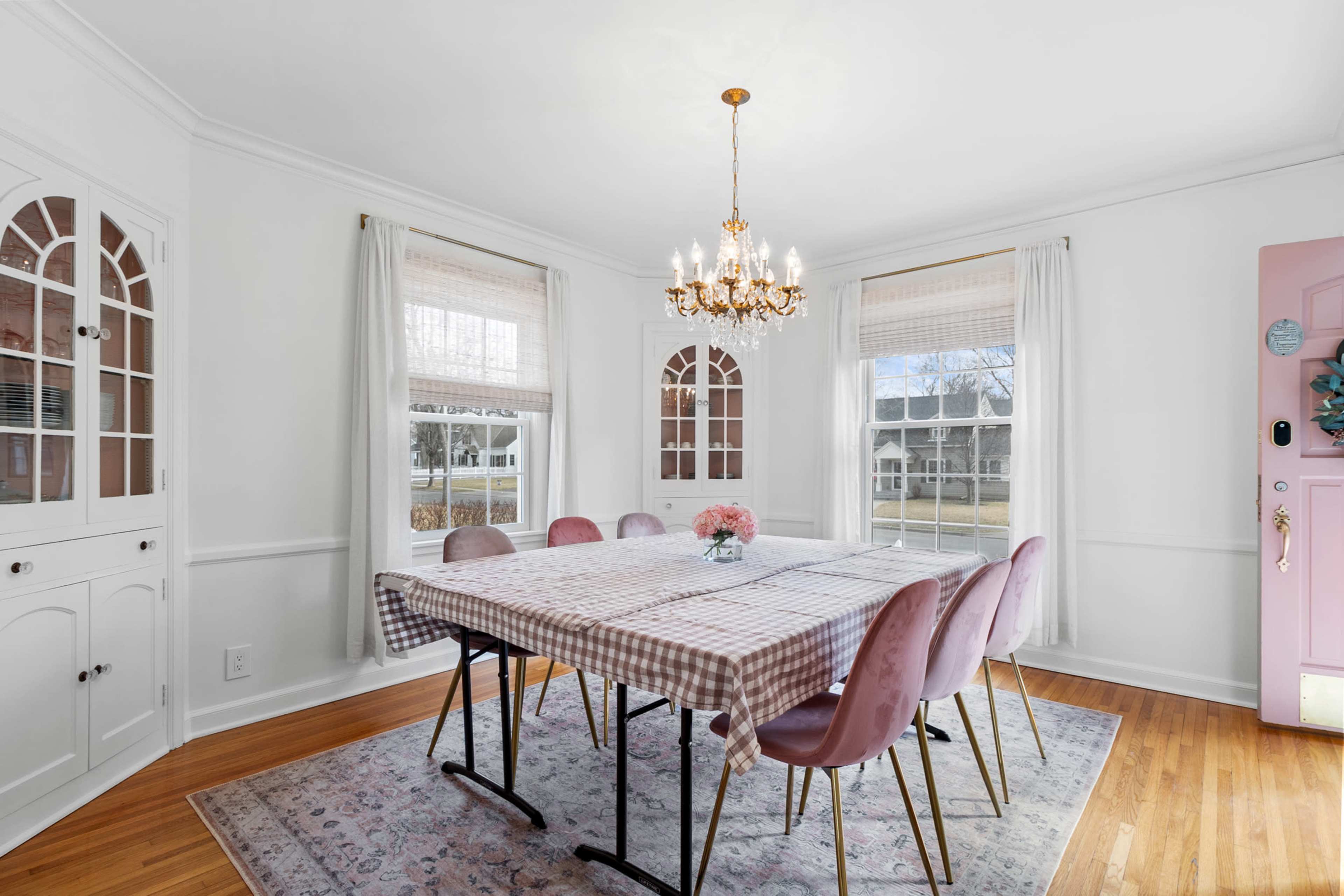 A dining room features a large checkered table surrounded by pink chairs, with a chandelier hanging above and large windows allowing natural light.