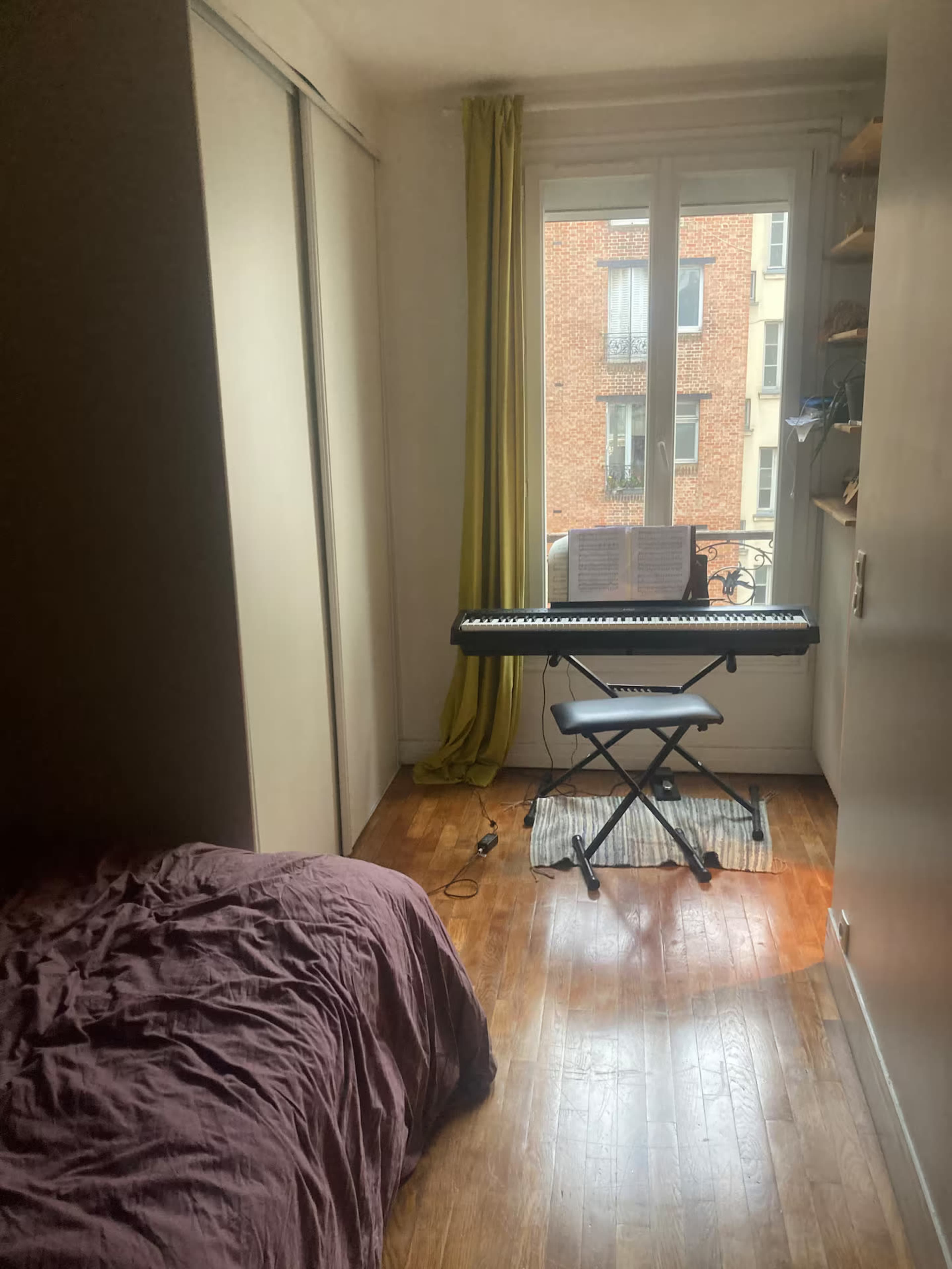 The image shows a small room with a bed on one side and an electronic keyboard set up near a window with green curtains.