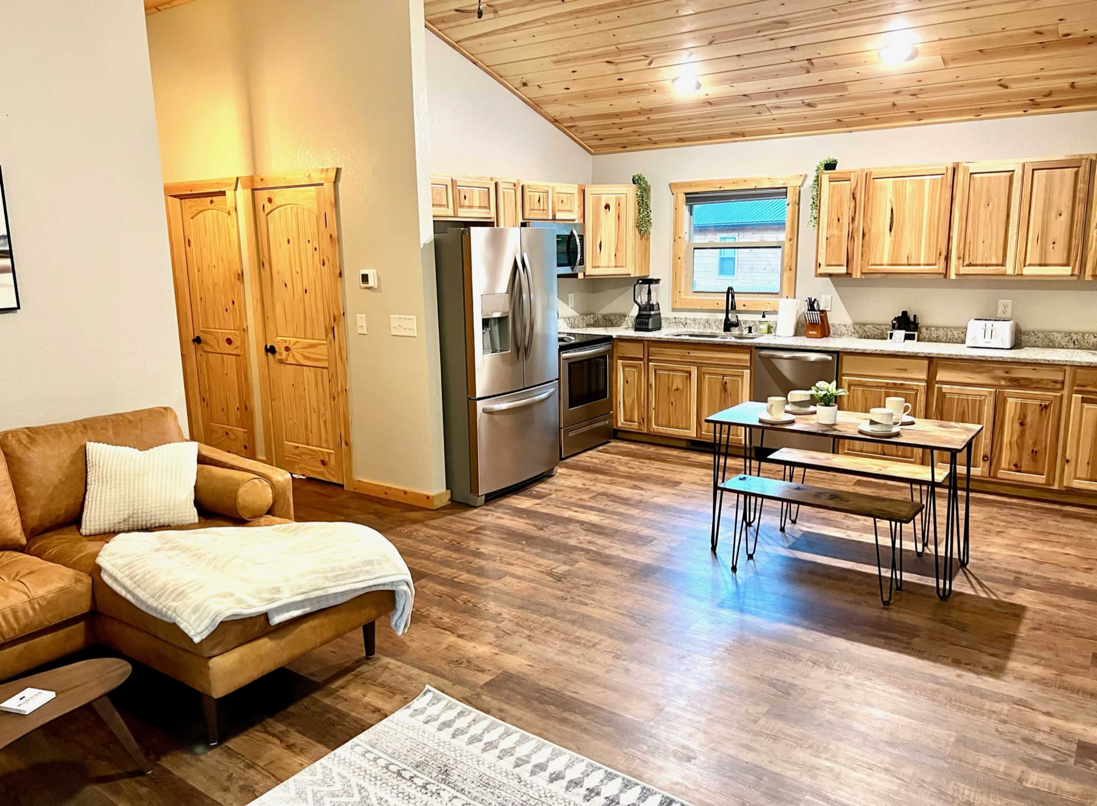 A modern kitchen and living area with wooden cabinetry, stainless steel appliances, a dining table set for four, and a cozy seating area featuring a brown leather couch.