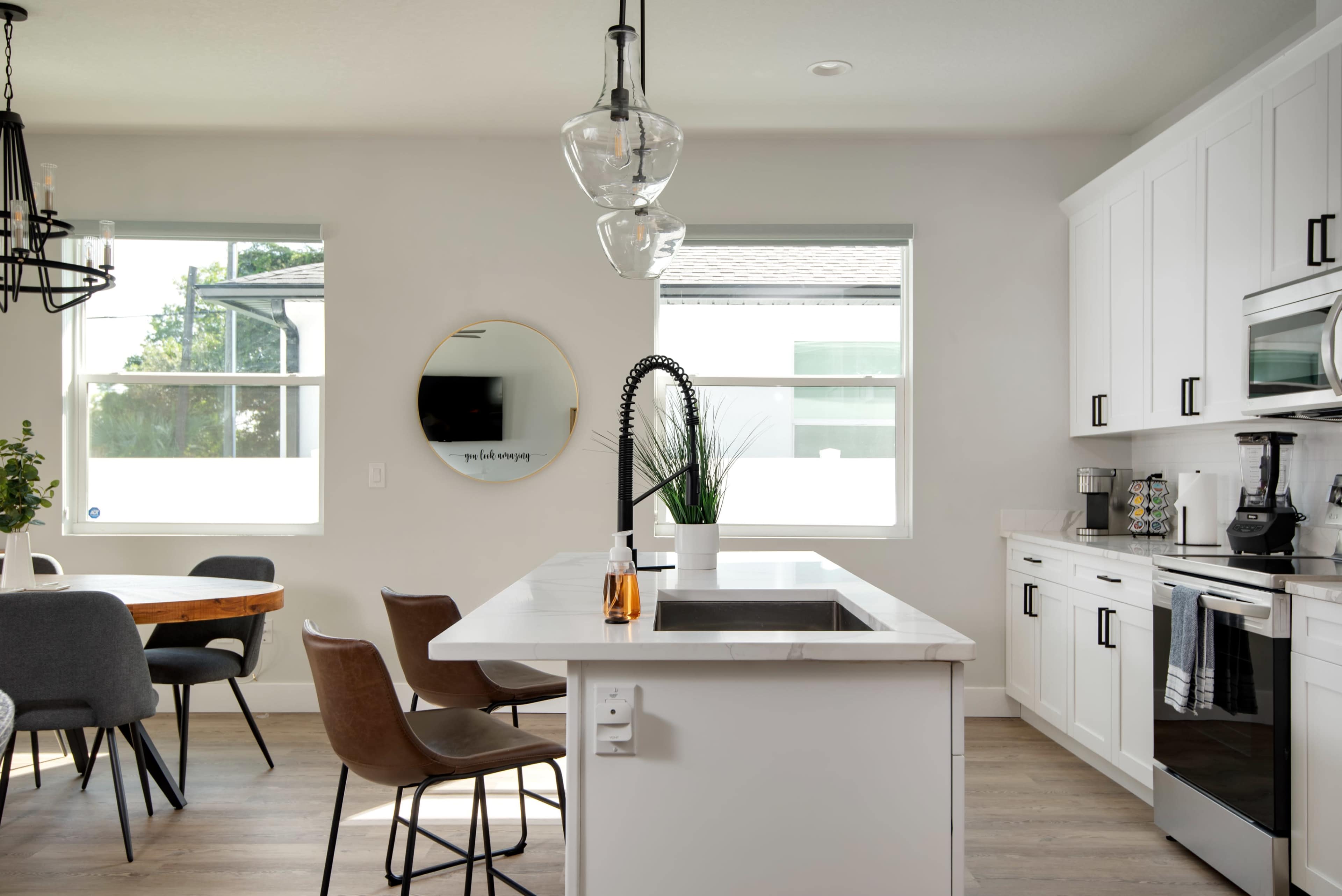 A modern kitchen features white cabinetry, a central island with a sink, a round dining table, and large windows providing natural light.