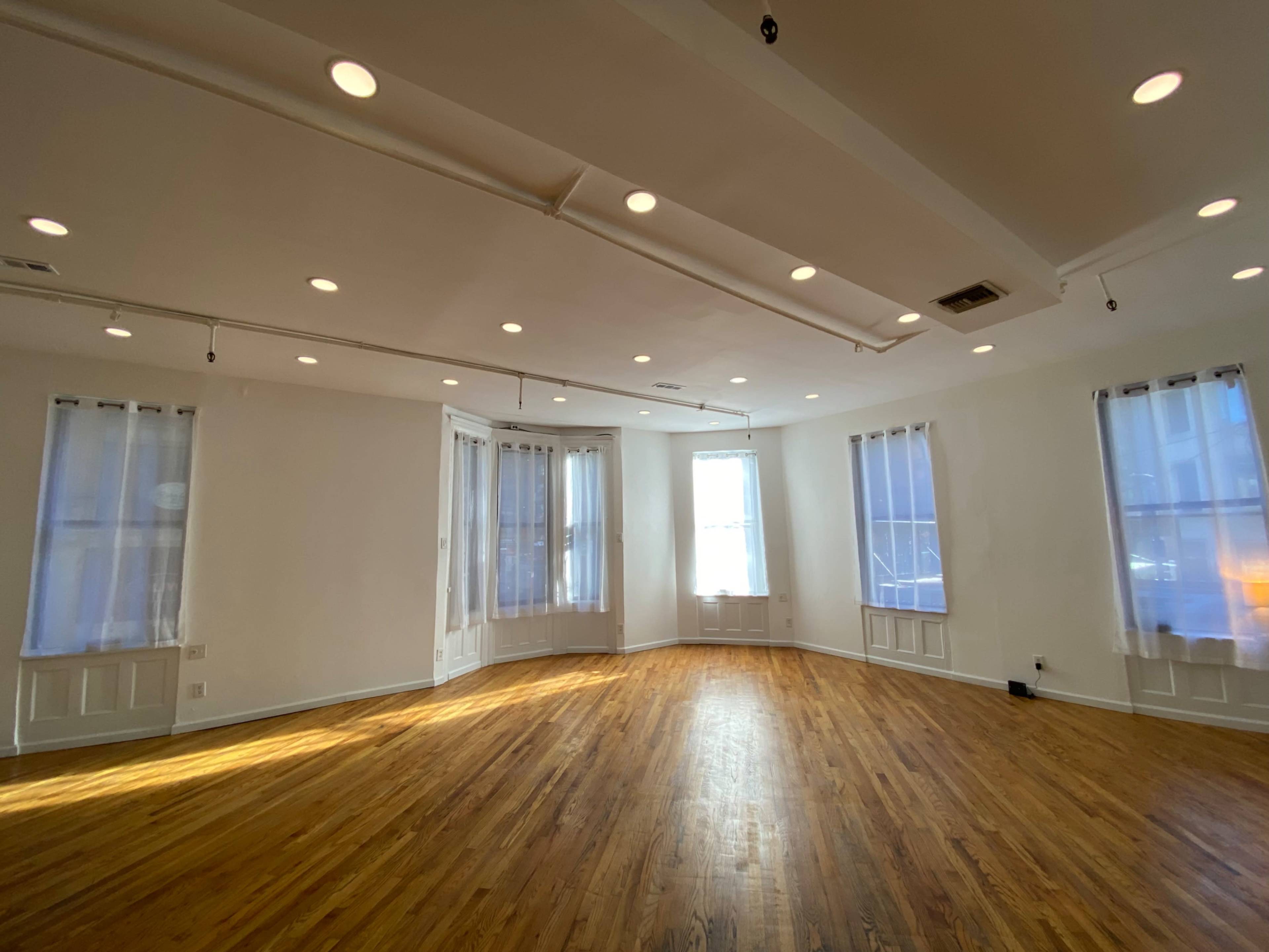 A spacious, well-lit room features wooden floors and large windows with sheer curtains.