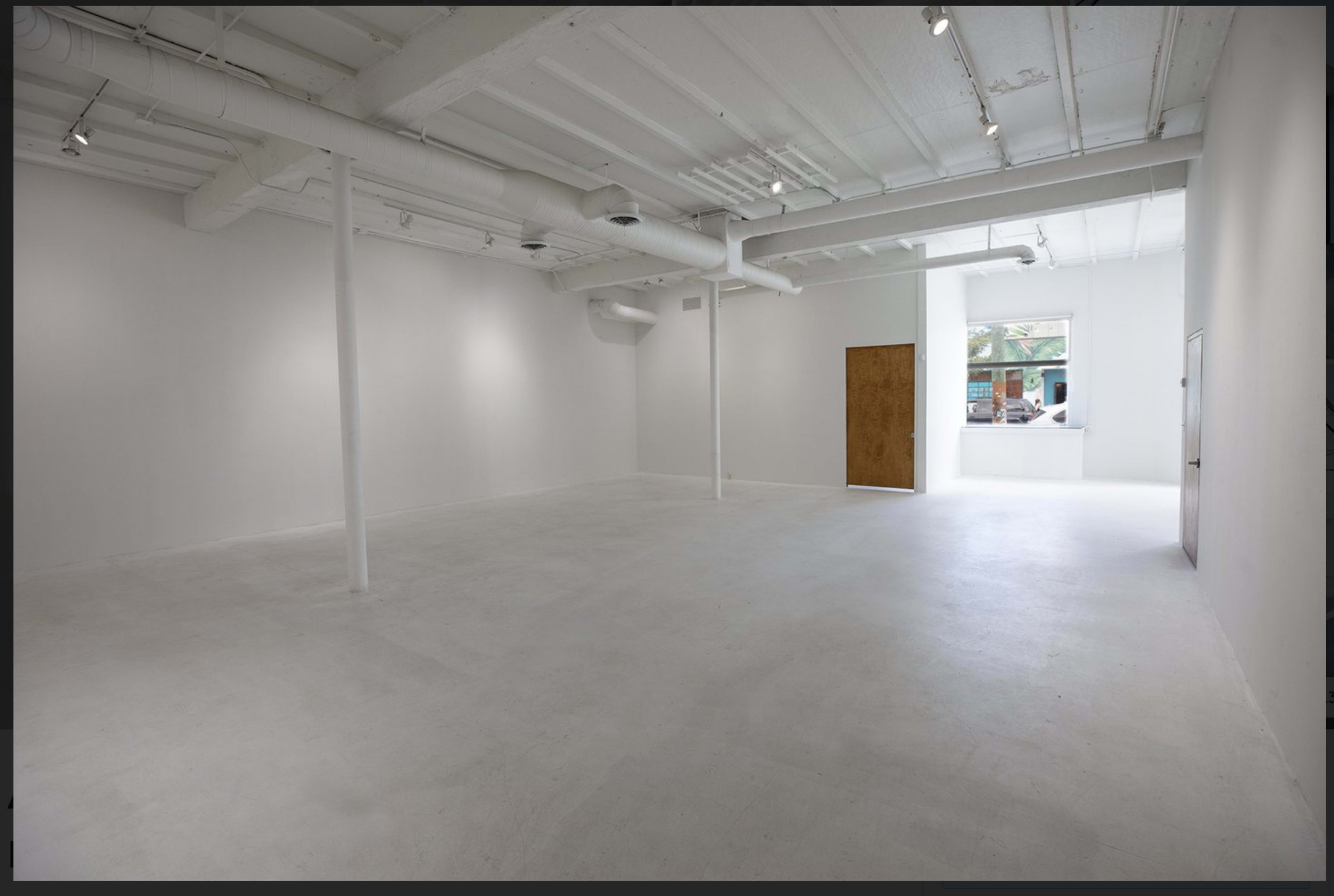 The image shows a spacious, empty white room with exposed beams and a large window facing an outdoor area.
