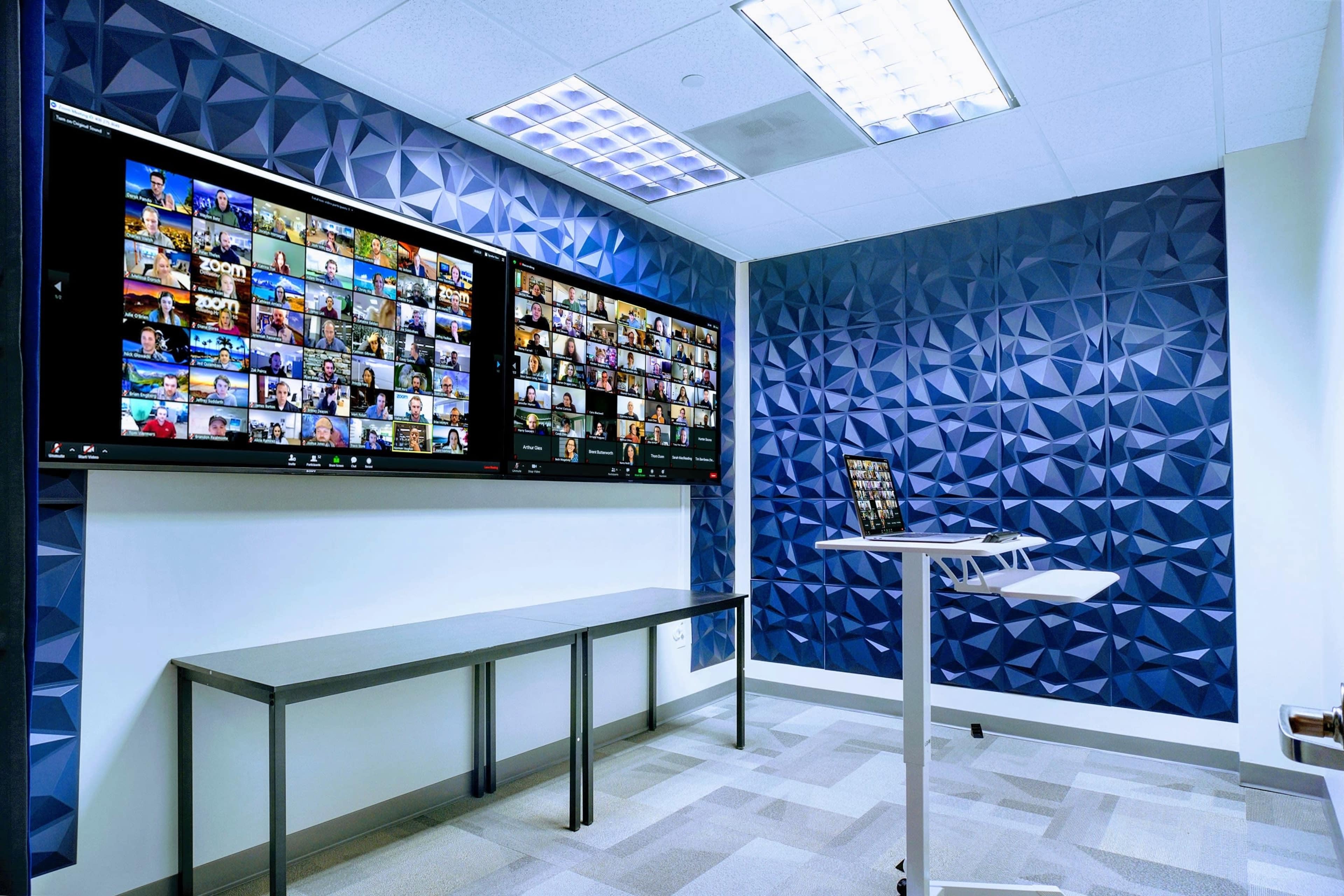 A modern conference room with two large screens displaying a grid of video conference participants on the left wall, and a standing table with a laptop on the right.