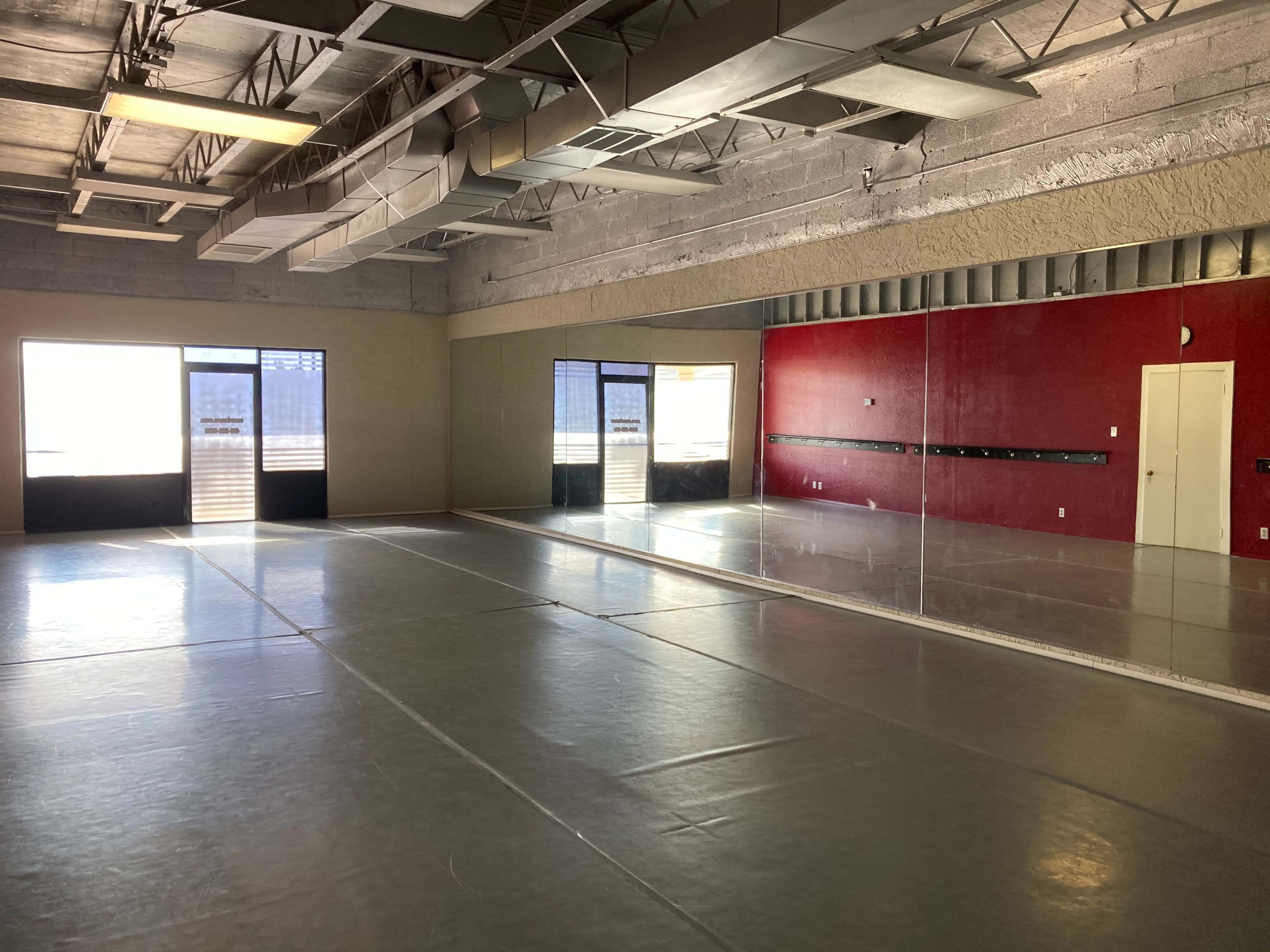 A spacious dance studio with large mirrors, wooden floors, and two sets of glass doors leading to an outside area.