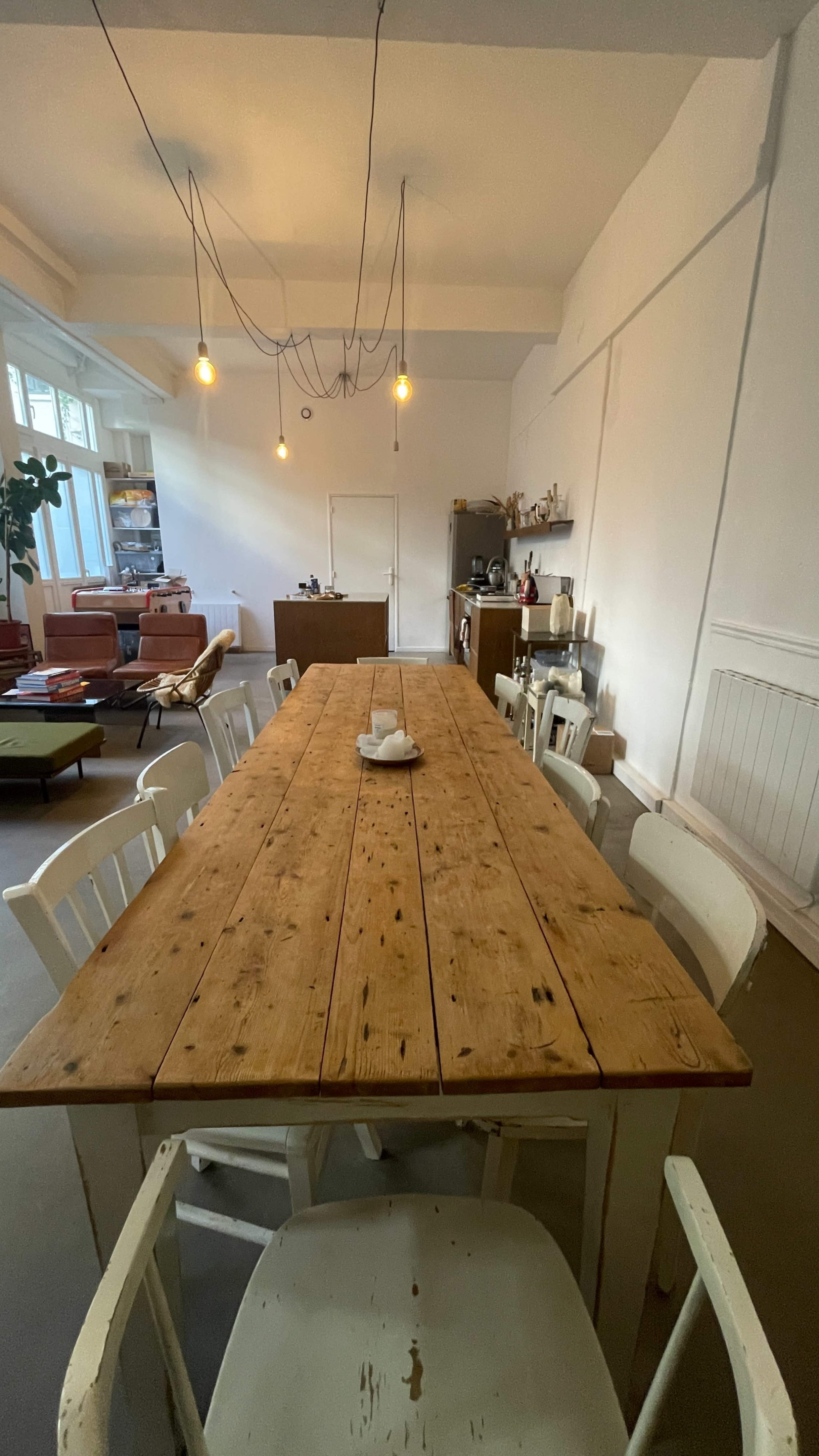 A long wooden dining table with white chairs is set in a well-lit room with minimal decor and exposed light bulbs hanging from the ceiling.