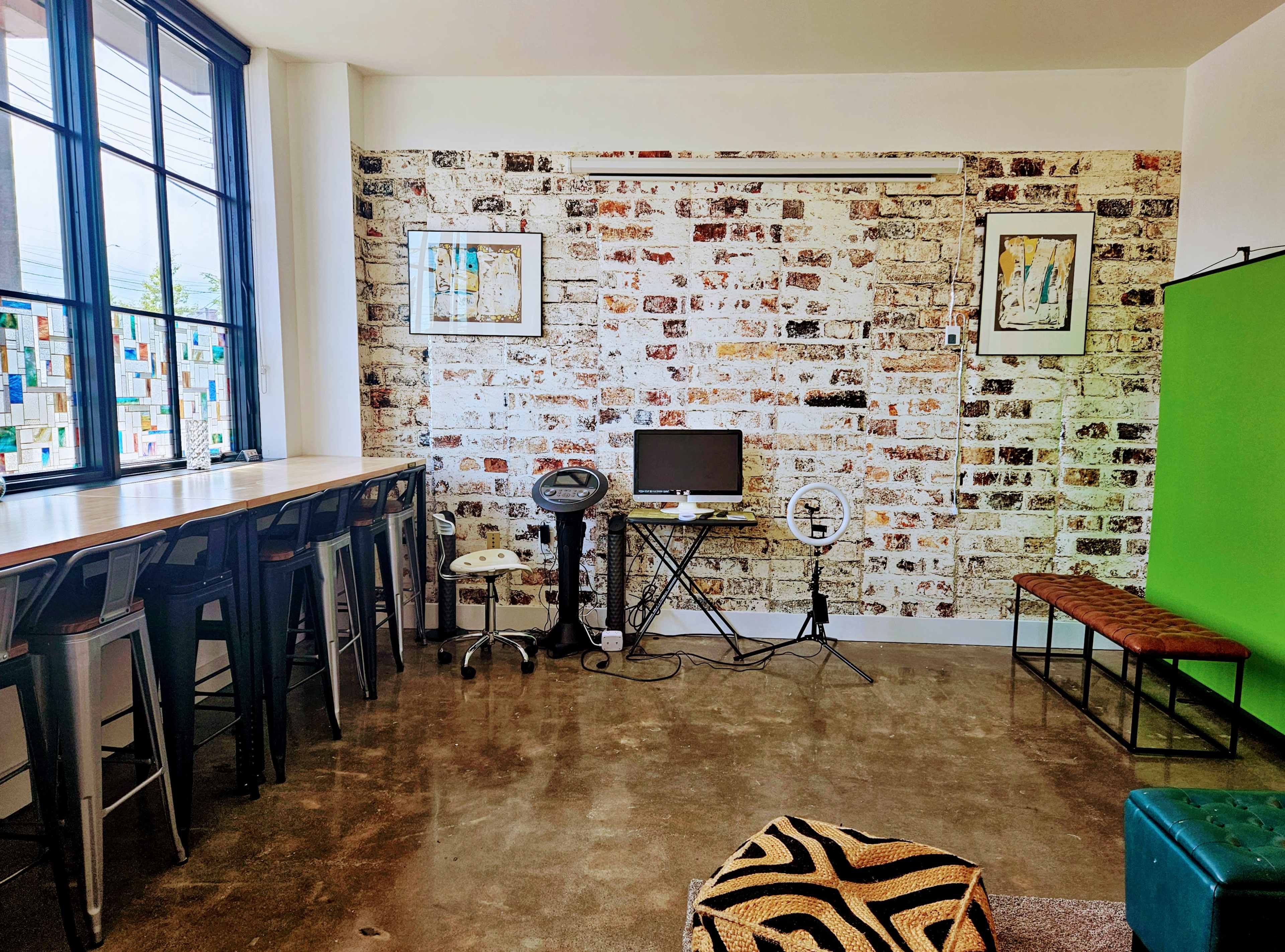 A room with a brick wall, a small desk with a monitor, a ring light on a stand, barstools along a countertop, and a green screen backdrop.