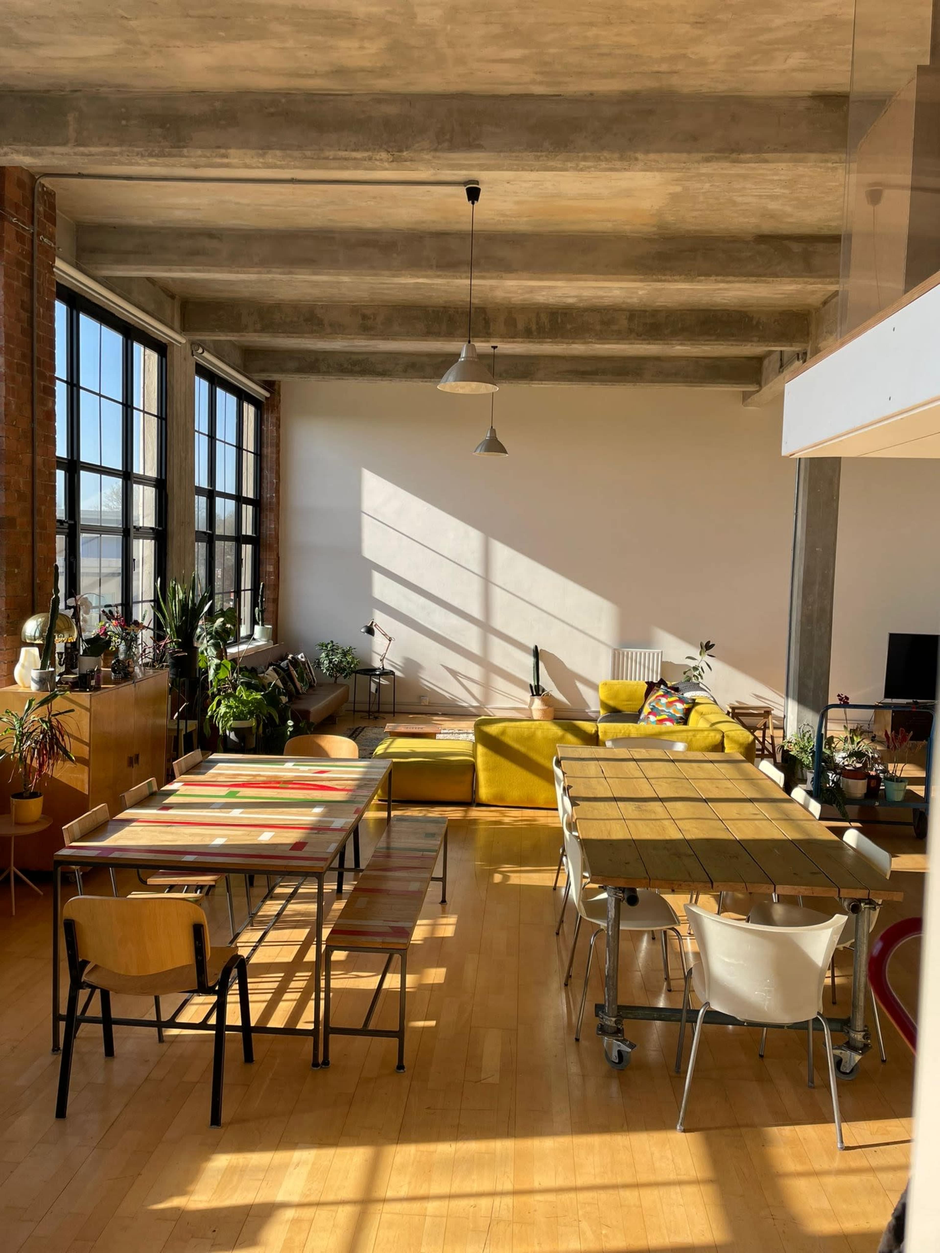 A spacious loft-style living area featuring yellow seating, multiple tables, large windows, and plants.