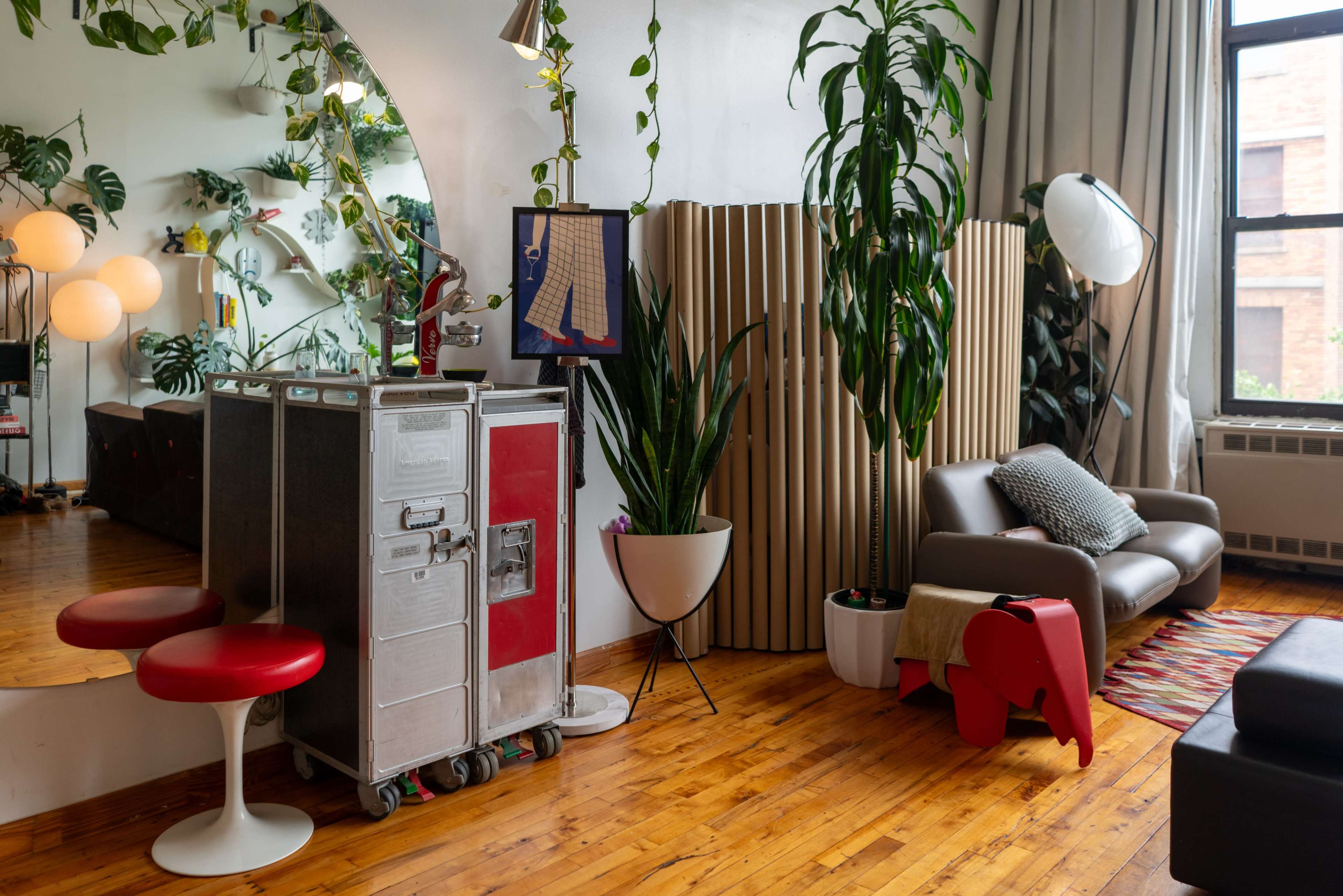 The image shows a stylish interior with wooden floors, a modern sofa, various plants, and a vintage cart along with a decorative room divider.