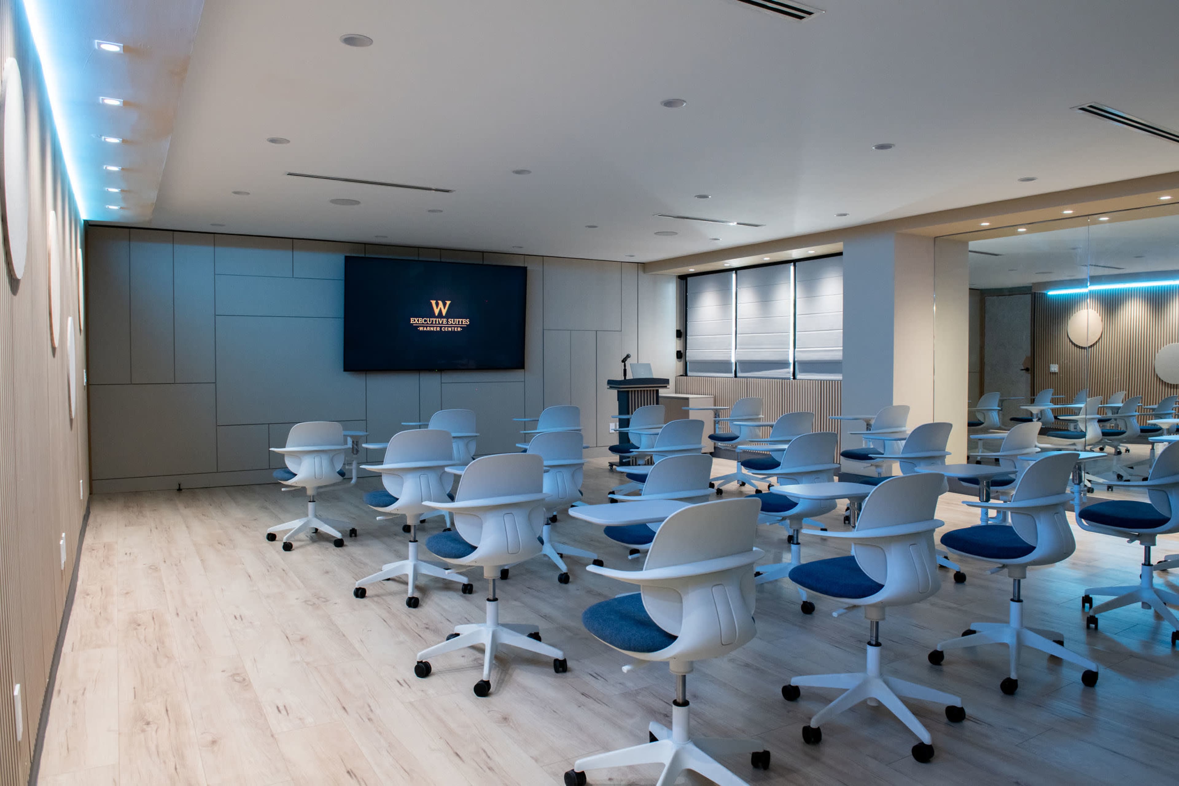 The image shows a modern conference room with rows of white rolling chairs facing a large screen on the front wall.