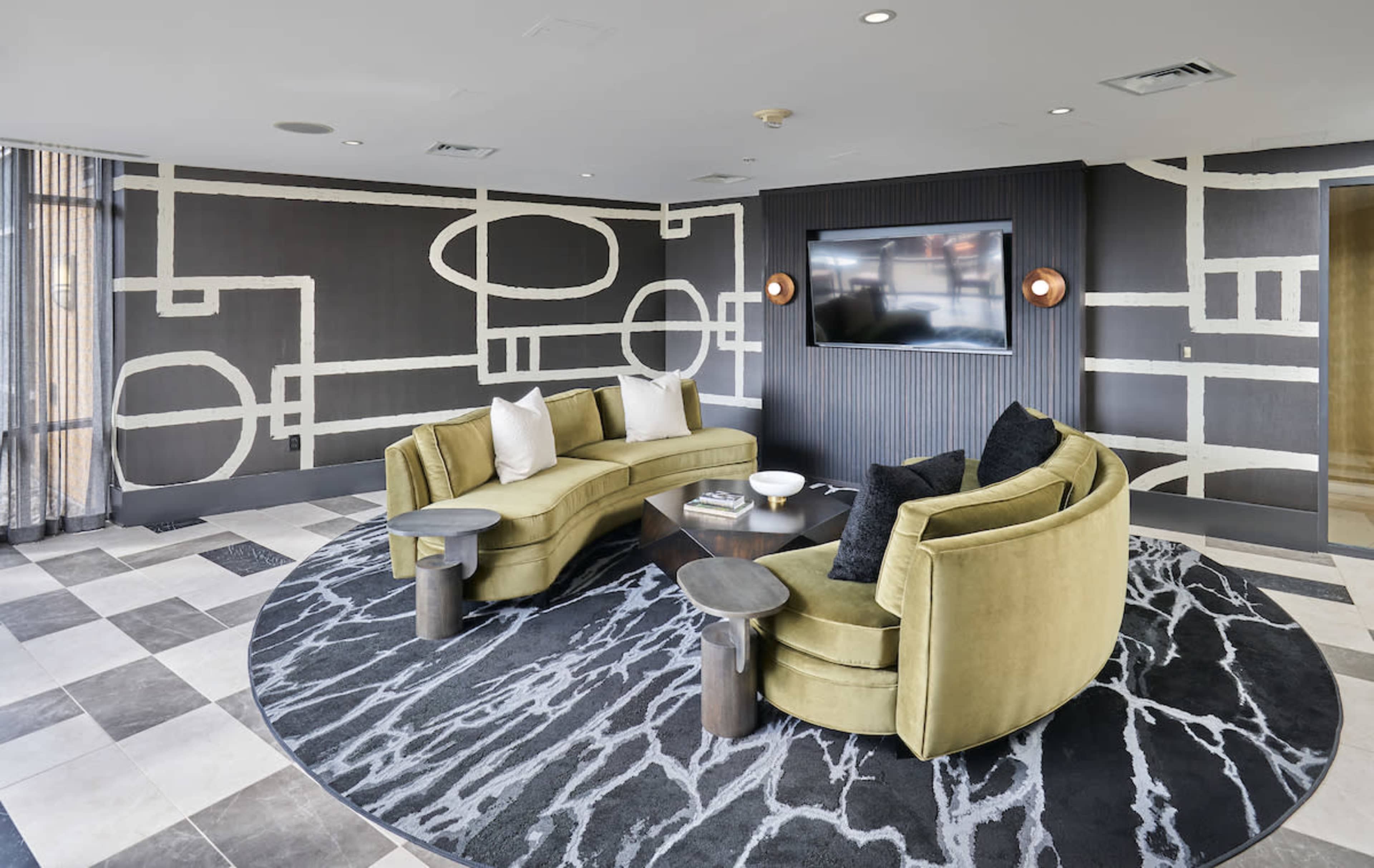 A modern lounge area features curved green sofas, a circular rug, and a large wall-mounted television.