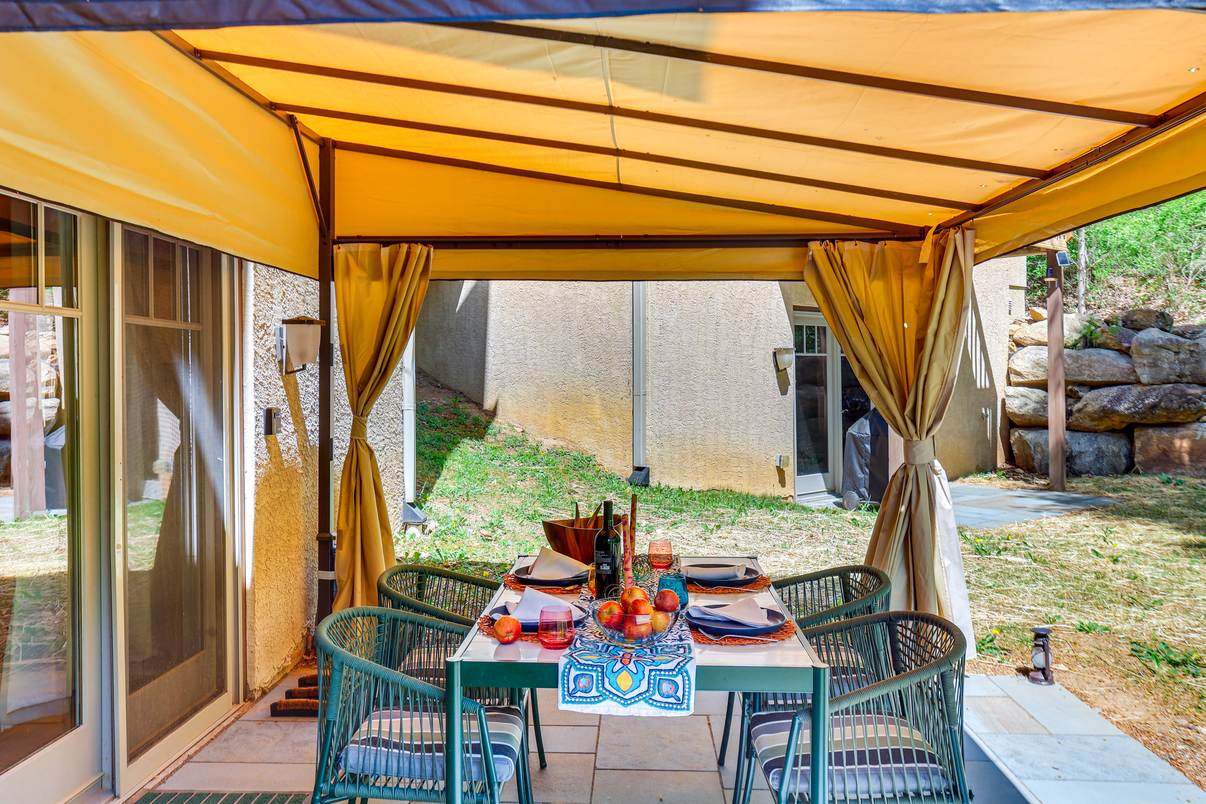 A patio area features a table set for dining under a yellow canopy, surrounded by green chairs and adorned with a colorful tablecloth and fruit.